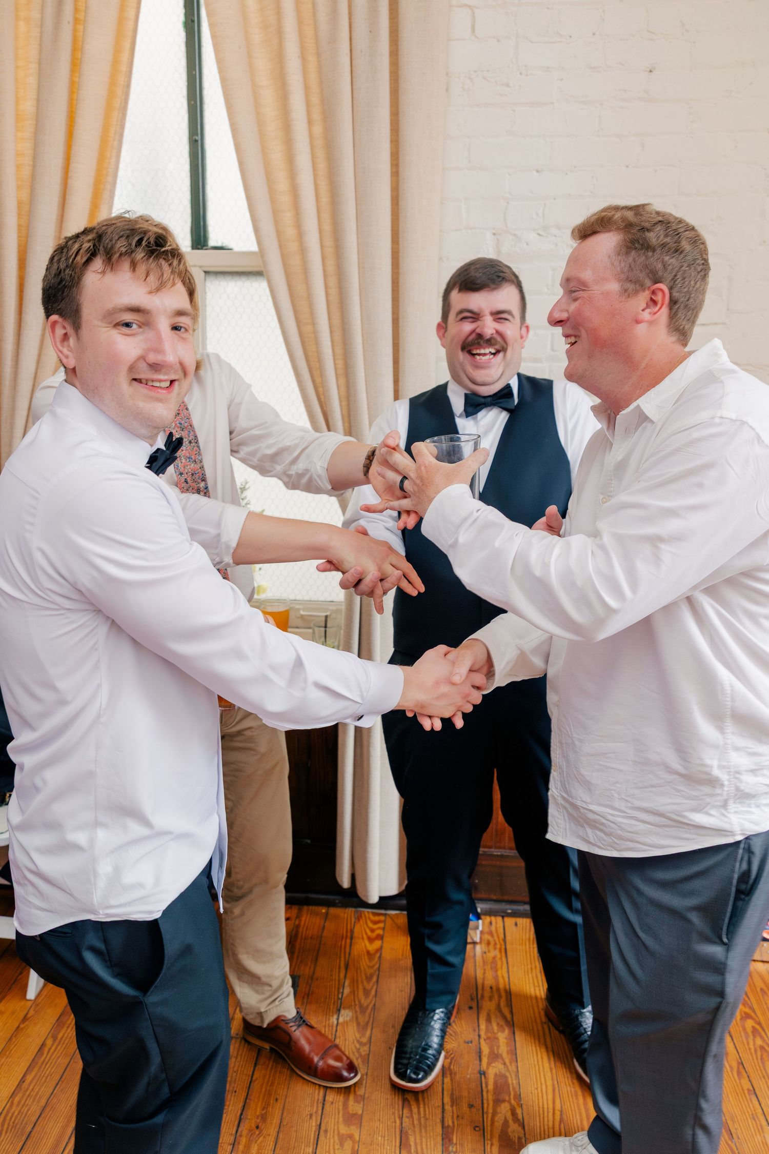 Three groomsmen in white shirts celebrate with handshakes and champagne toast at a wedding reception.