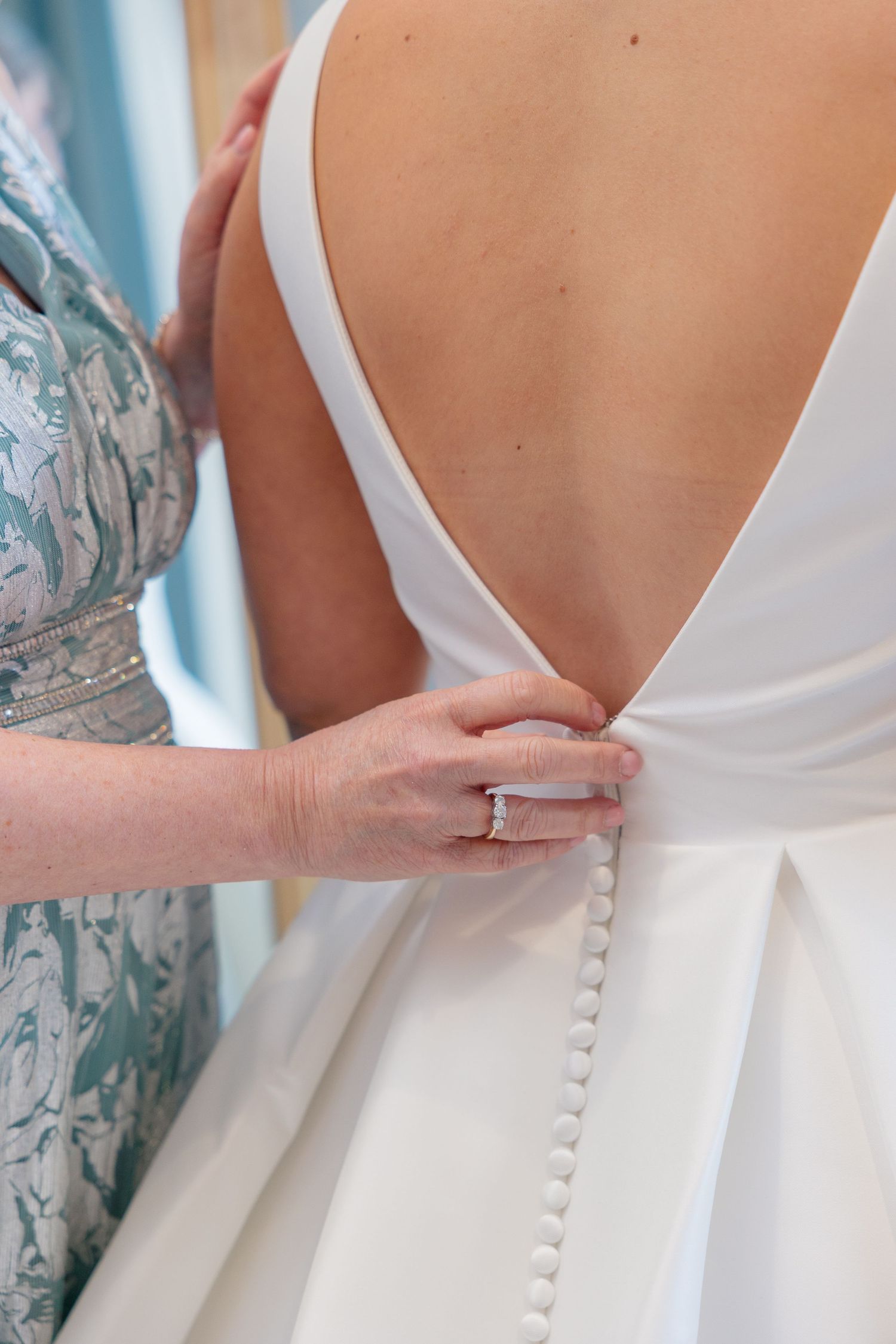 Close-up of bride's open back wedding gown with delicate button detail being fastened by a helper.