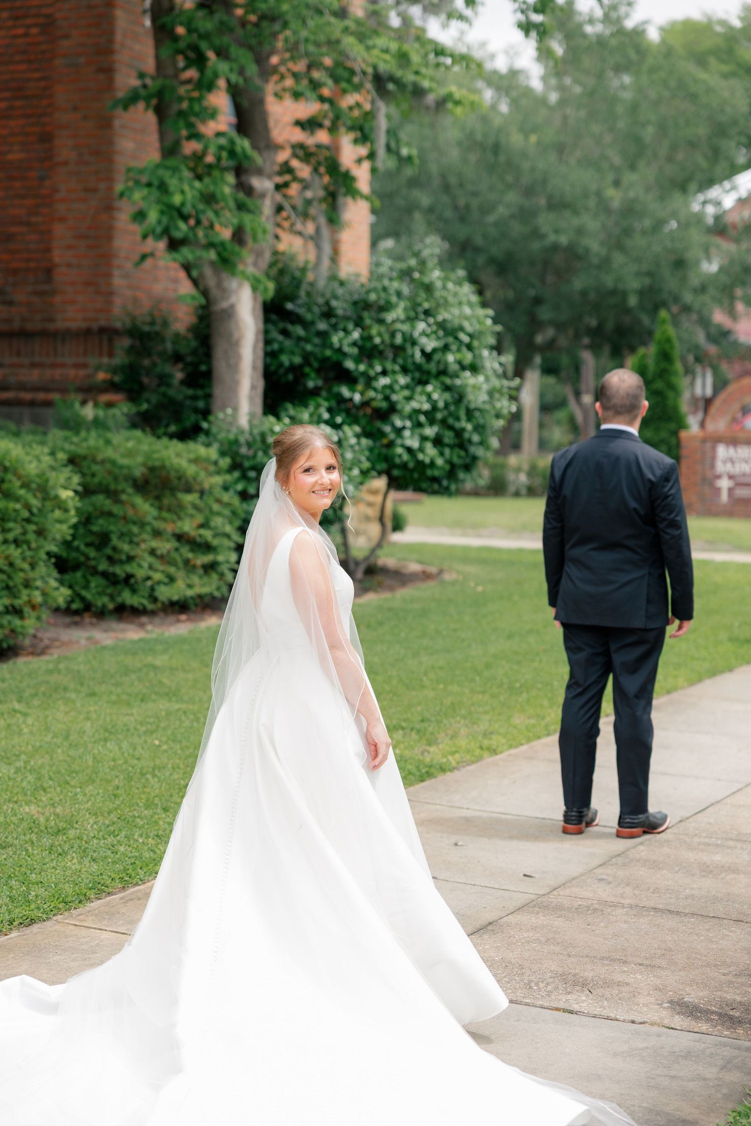 Bride in flowing white gown and veil glances back during first look with groom outside church.