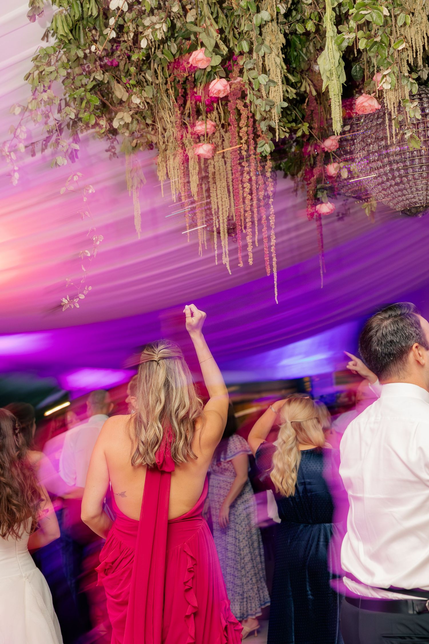 Wedding guests dance under a floral chandelier with purple lighting at a lively reception.