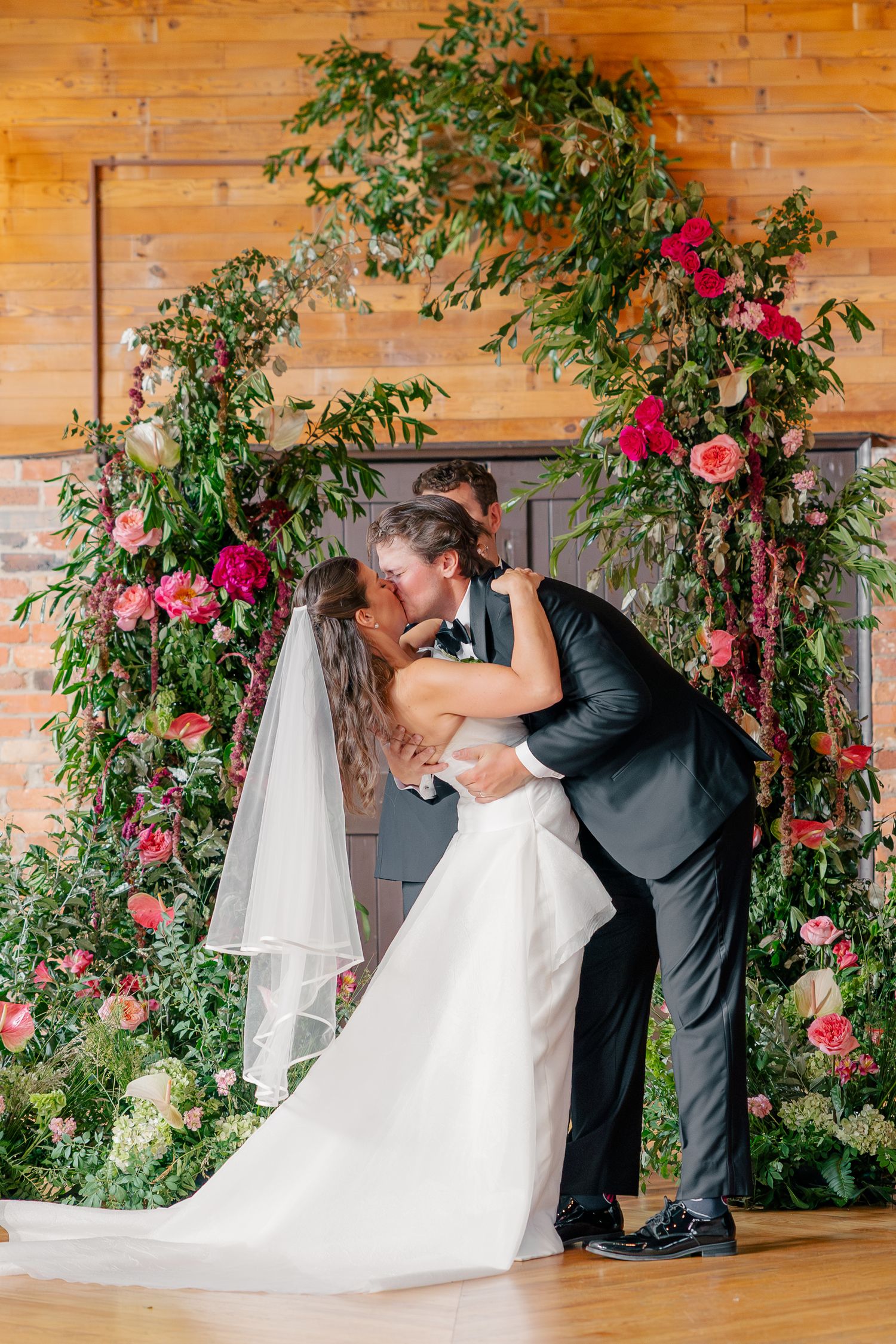 Bride and groom share first kiss under lush floral arch of pink and green blooms.