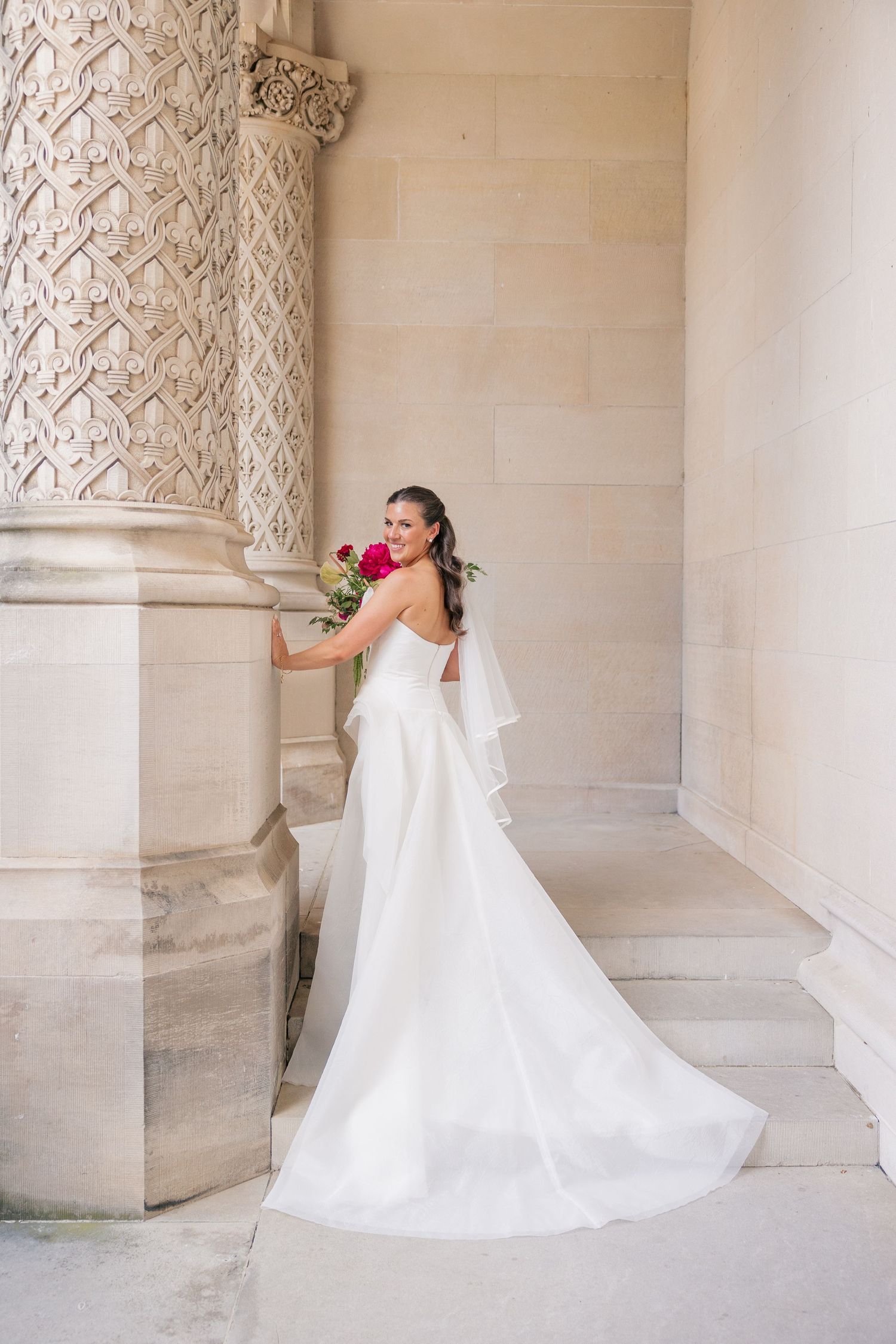Elegant bride in white gown with train poses beside ornate carved stone column.
