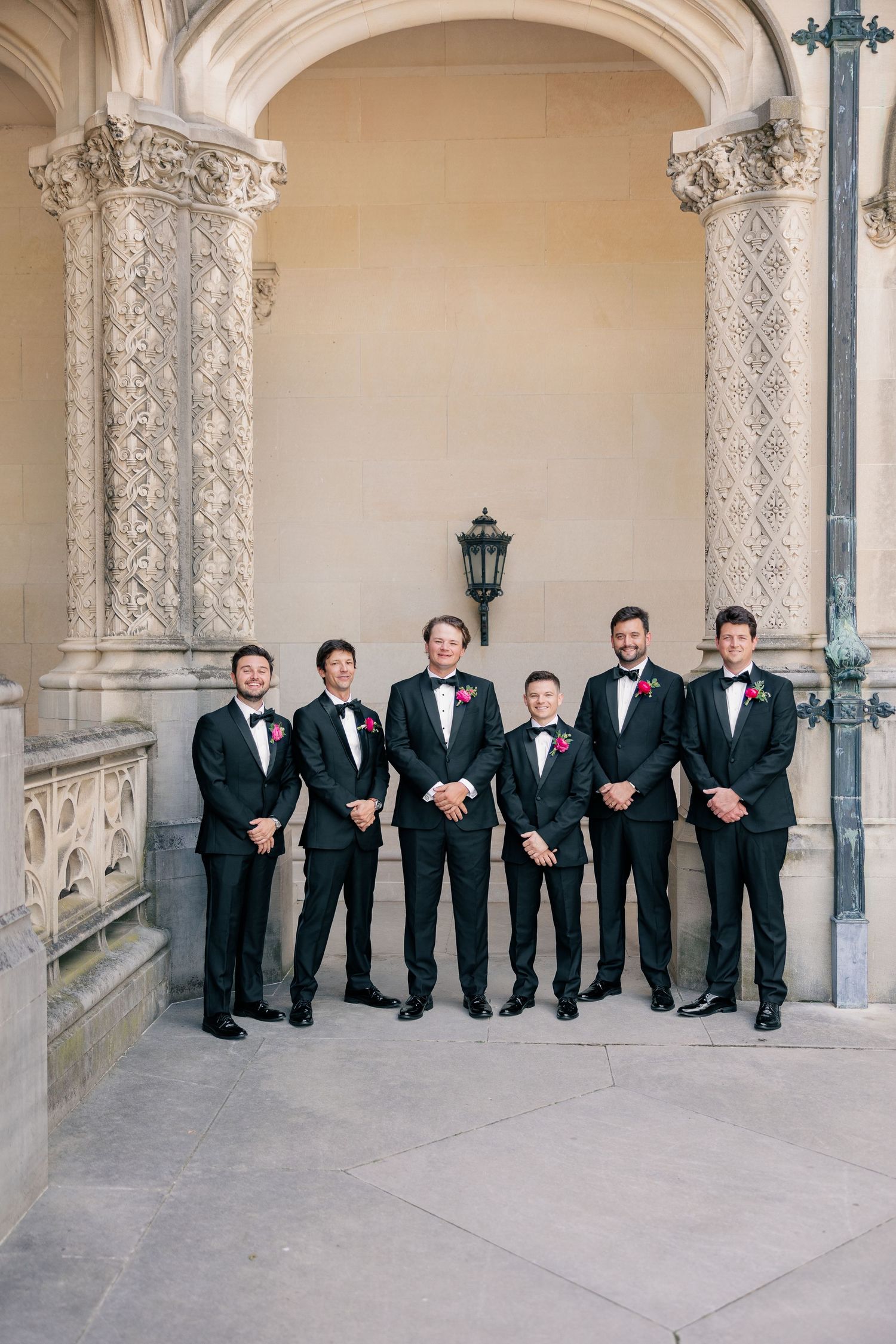 Six groomsmen in black tuxedos with pink boutonnieres stand beneath ornate stone archway.
