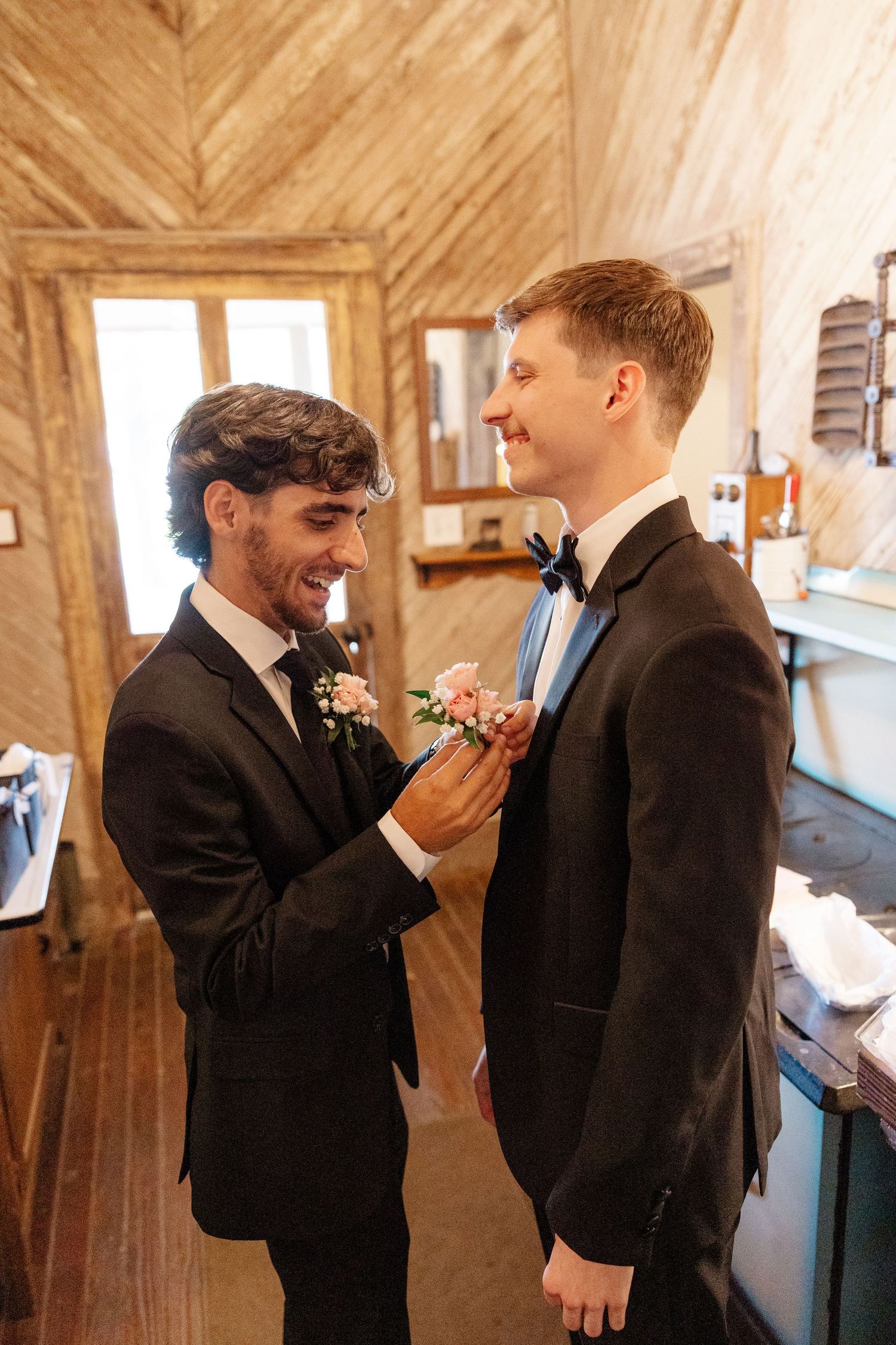 Two groomsmen share a lighthearted moment in a rustic wooden church interior.
