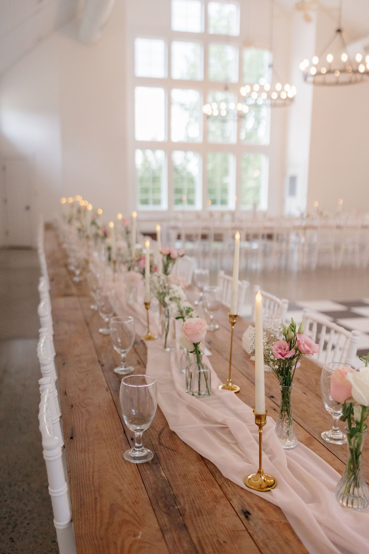 Long rustic wooden table set for wedding reception with pink runner and delicate floral centerpieces.