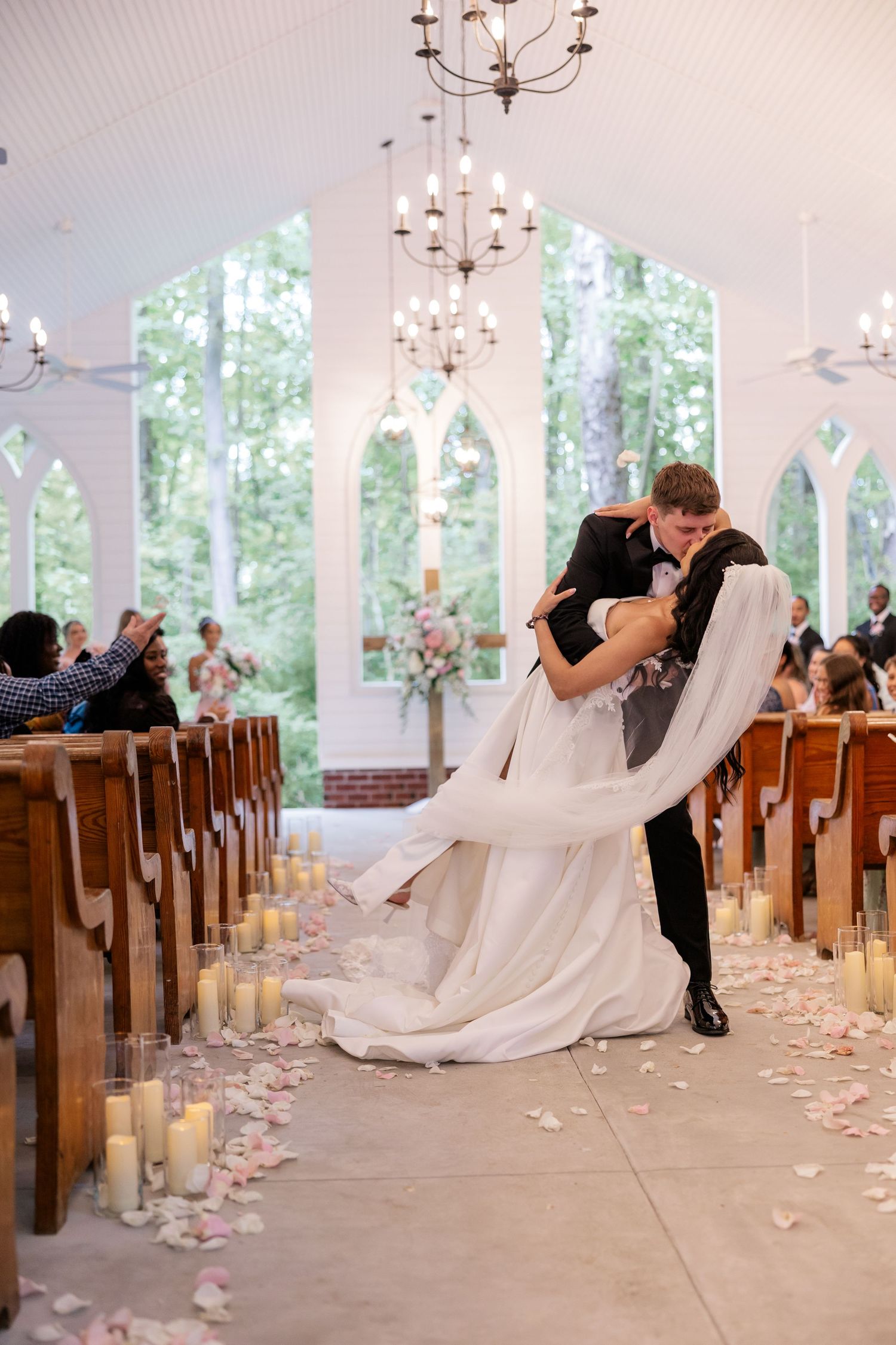 Wedding couple dipping and kissing in a chapel aisle lined with wooden pews and glowing candles.