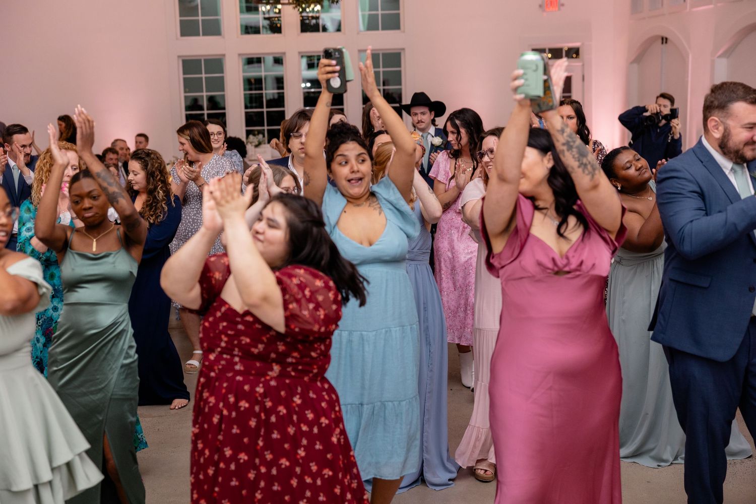 Wedding reception celebration with guests in colorful attire dancing and cheering in a pink-walled venue.
