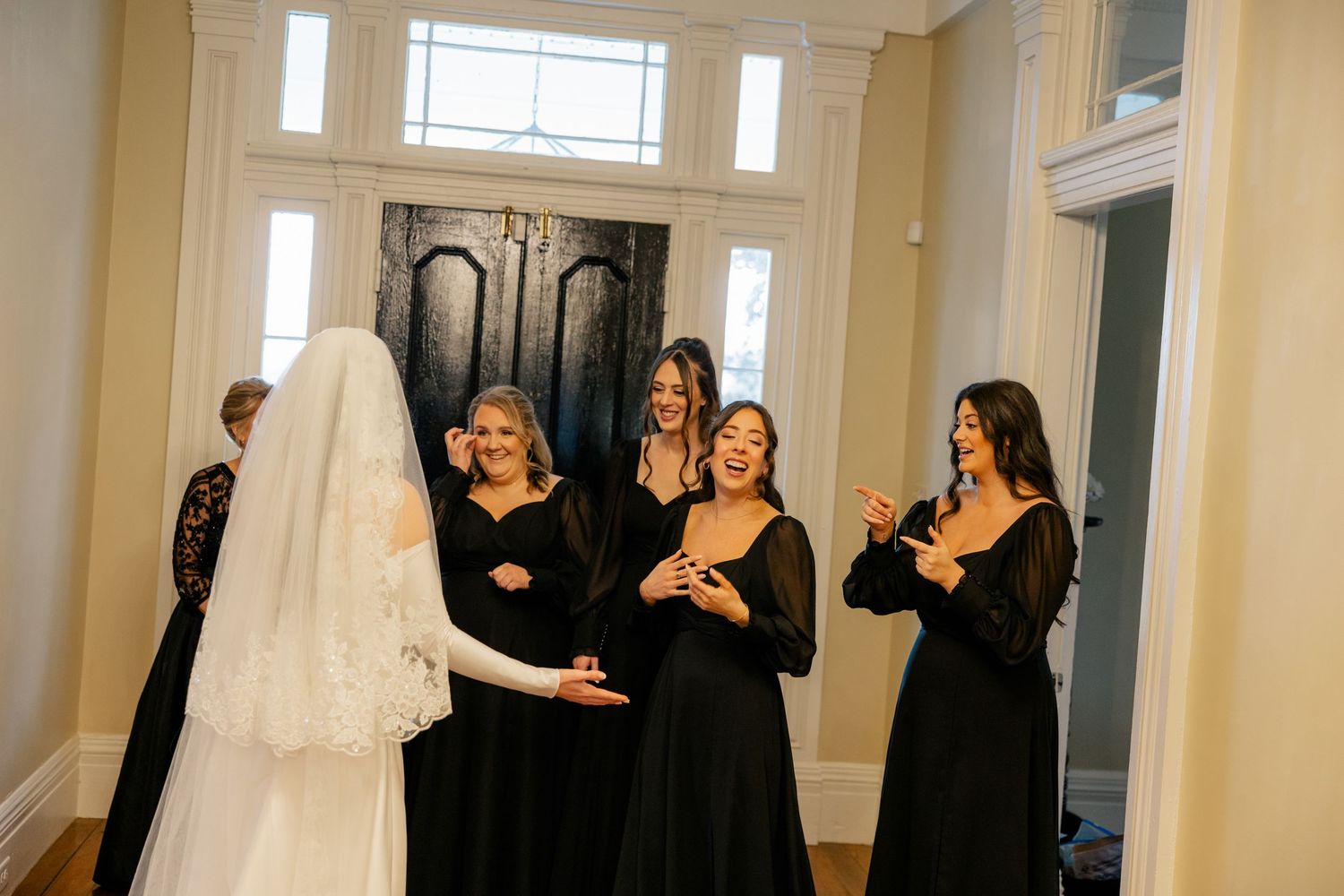 Wedding party in black dresses gathers together in brightly lit room with tall windows.