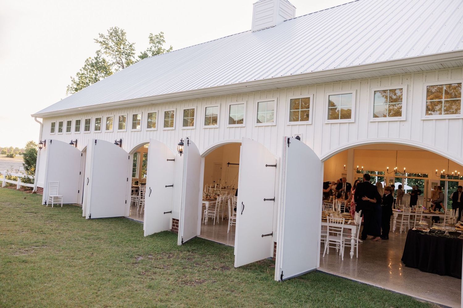 Large white barn-style wedding venue with multiple open doors revealing elegant indoor reception setup.