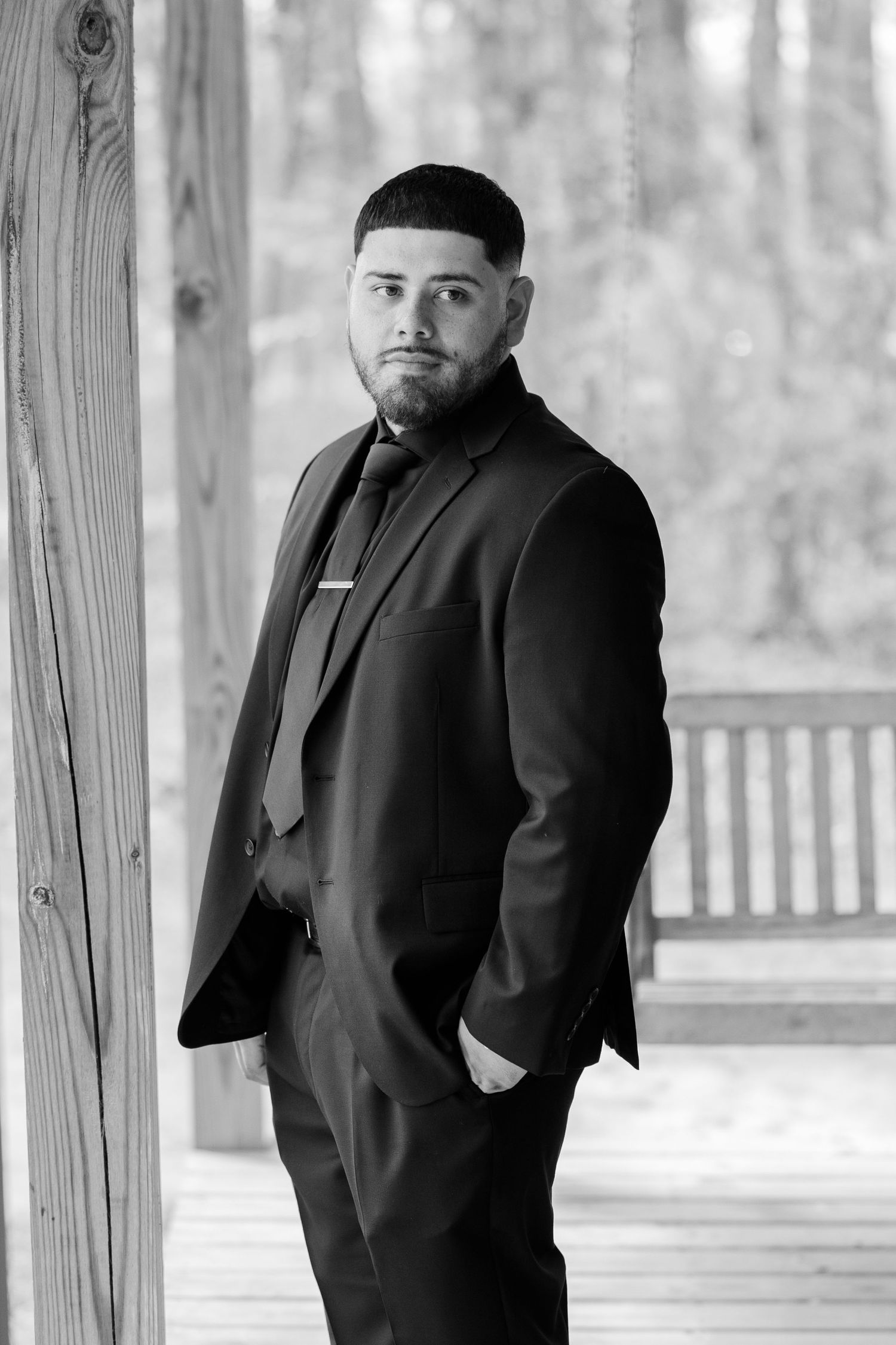 A black and white portrait of someone in formal attire leaning against a wooden post.