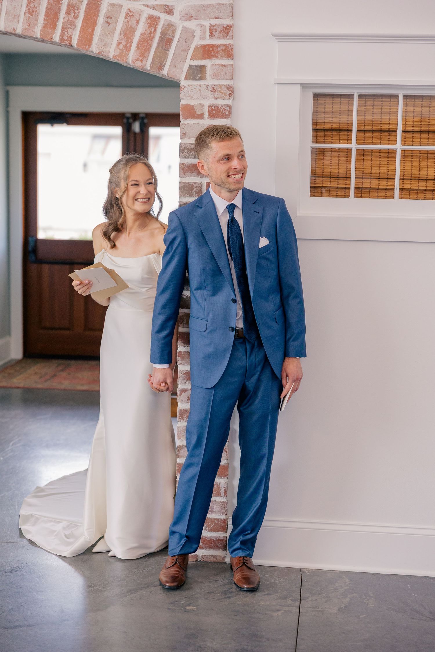 Bride in white gown reads vows while groom in blue suit smiles during wedding first look indoors.