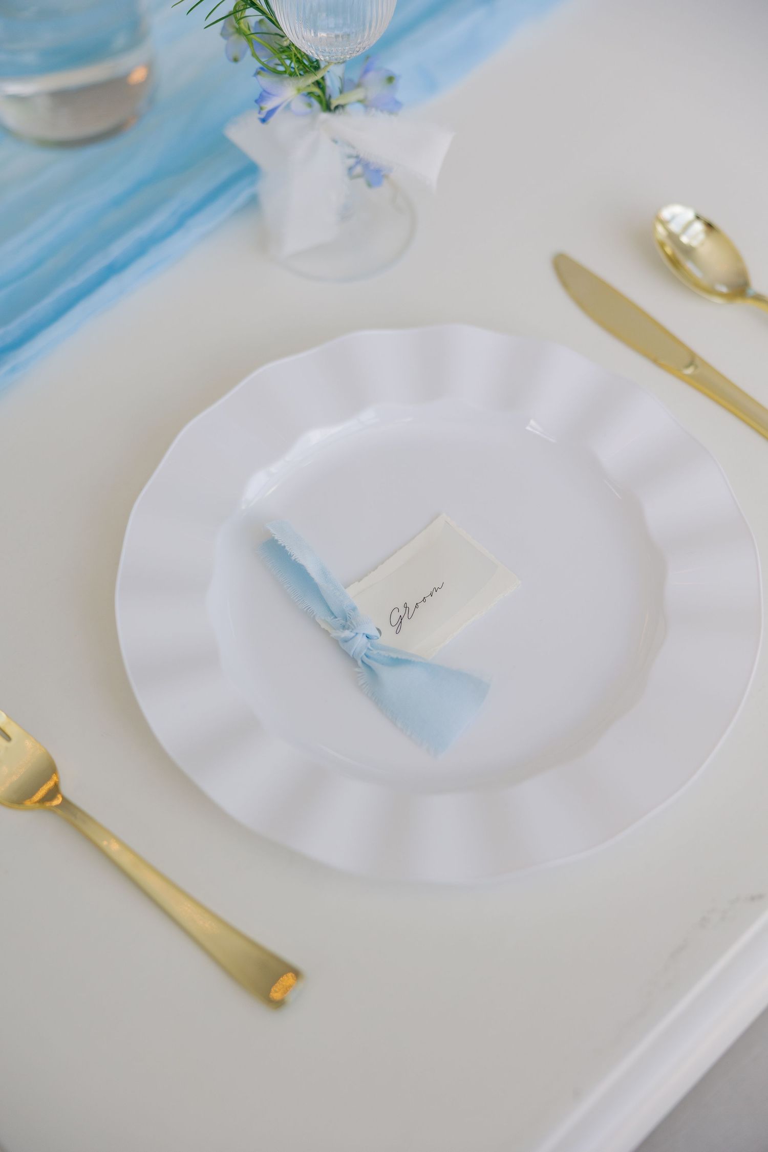 Elegant white scalloped plate with blue ribbon place card and gold cutlery on white wedding reception table.