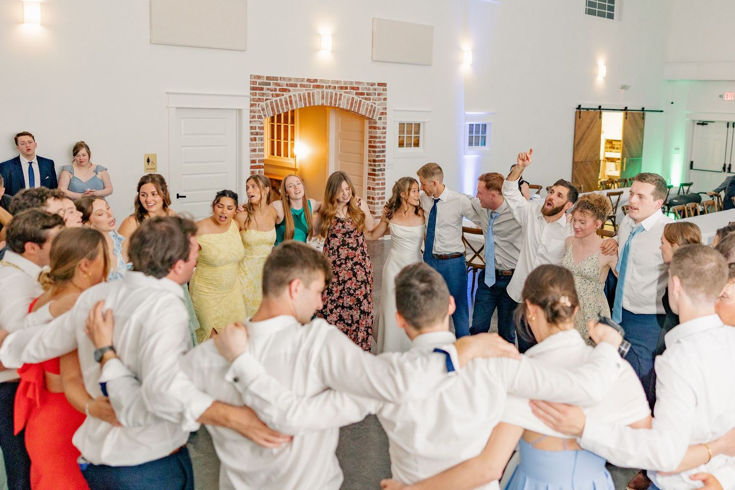 Wedding guests form a large circle dance on the floor of a bright white reception hall.