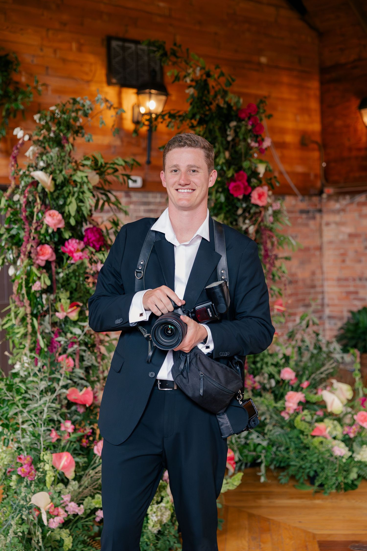 Smiling male wedding photographer in navy suit holding camera, standing before lush floral arch with pink roses at elegant venue.
