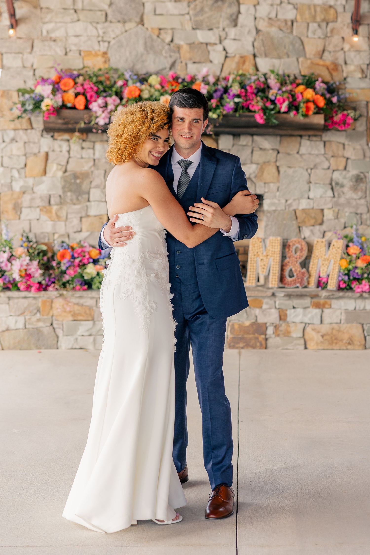 Bride and groom embrace smiling in front of a stone wall with colorful flowers and M&M marquee letters.