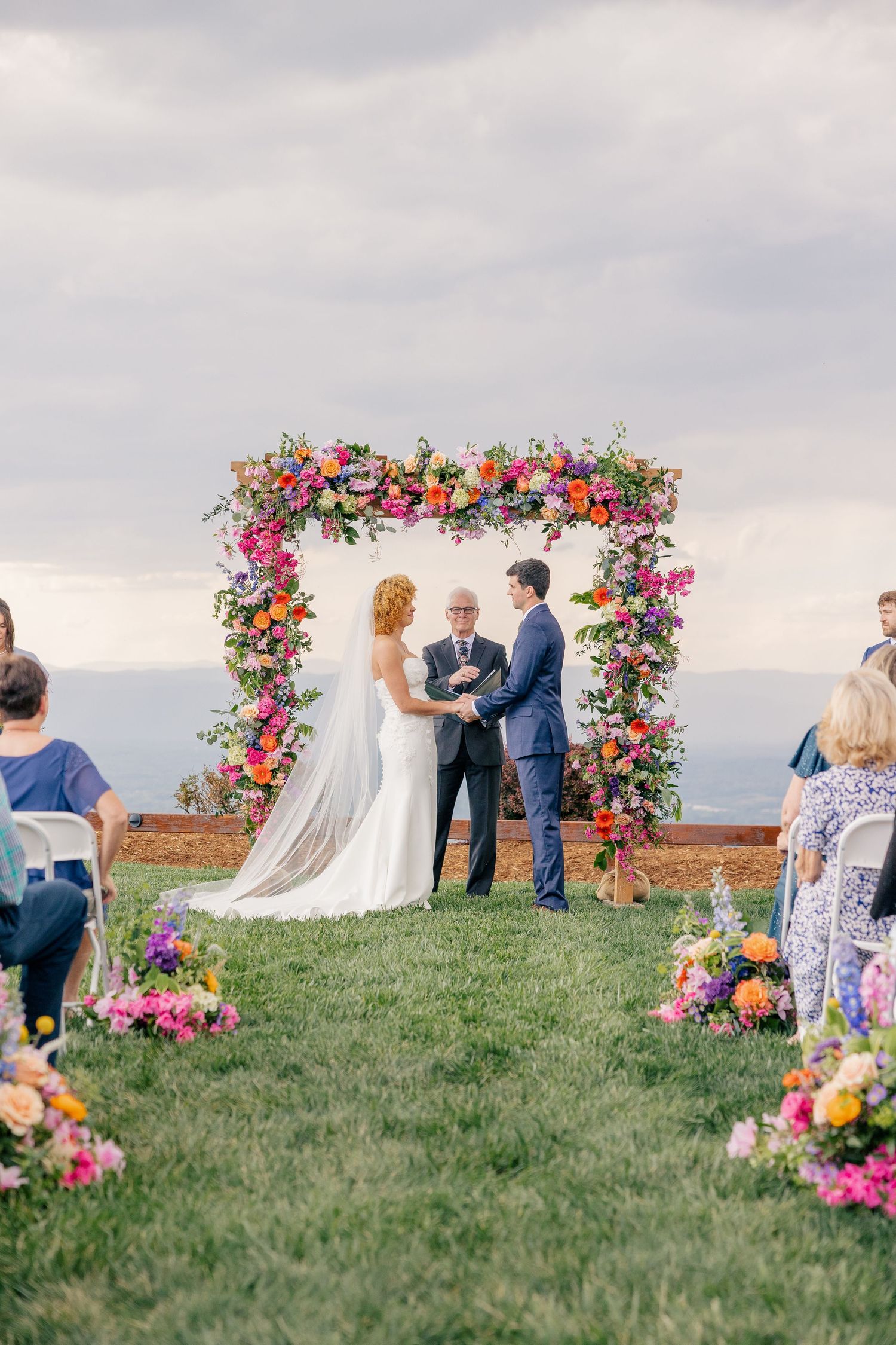 Bride and groom exchange vows under a vibrant floral arch at an outdoor wedding ceremony with scenic views.