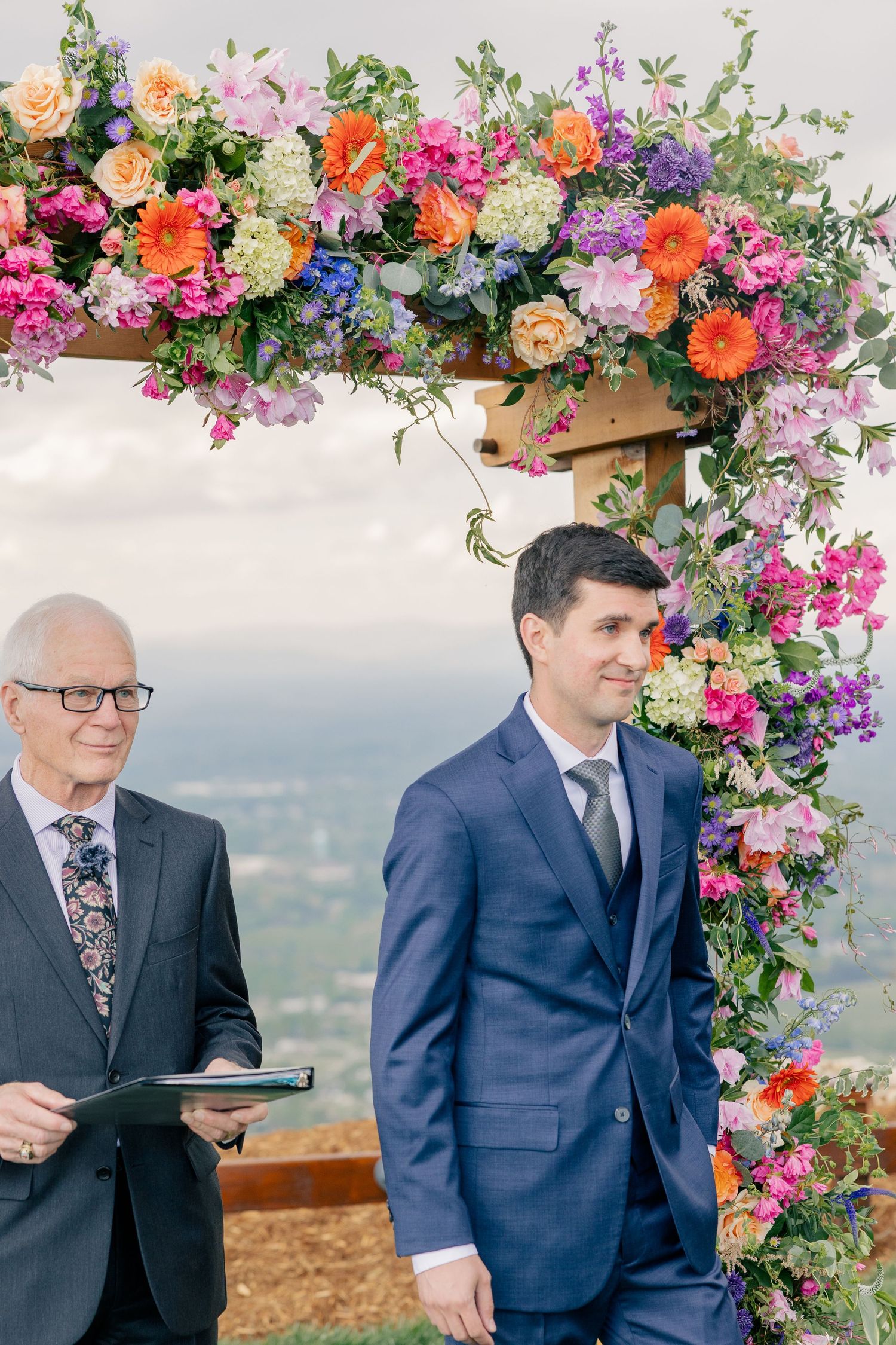 Groom in navy suit stands with officiant under a lush colorful floral wedding arch with panoramic views.
