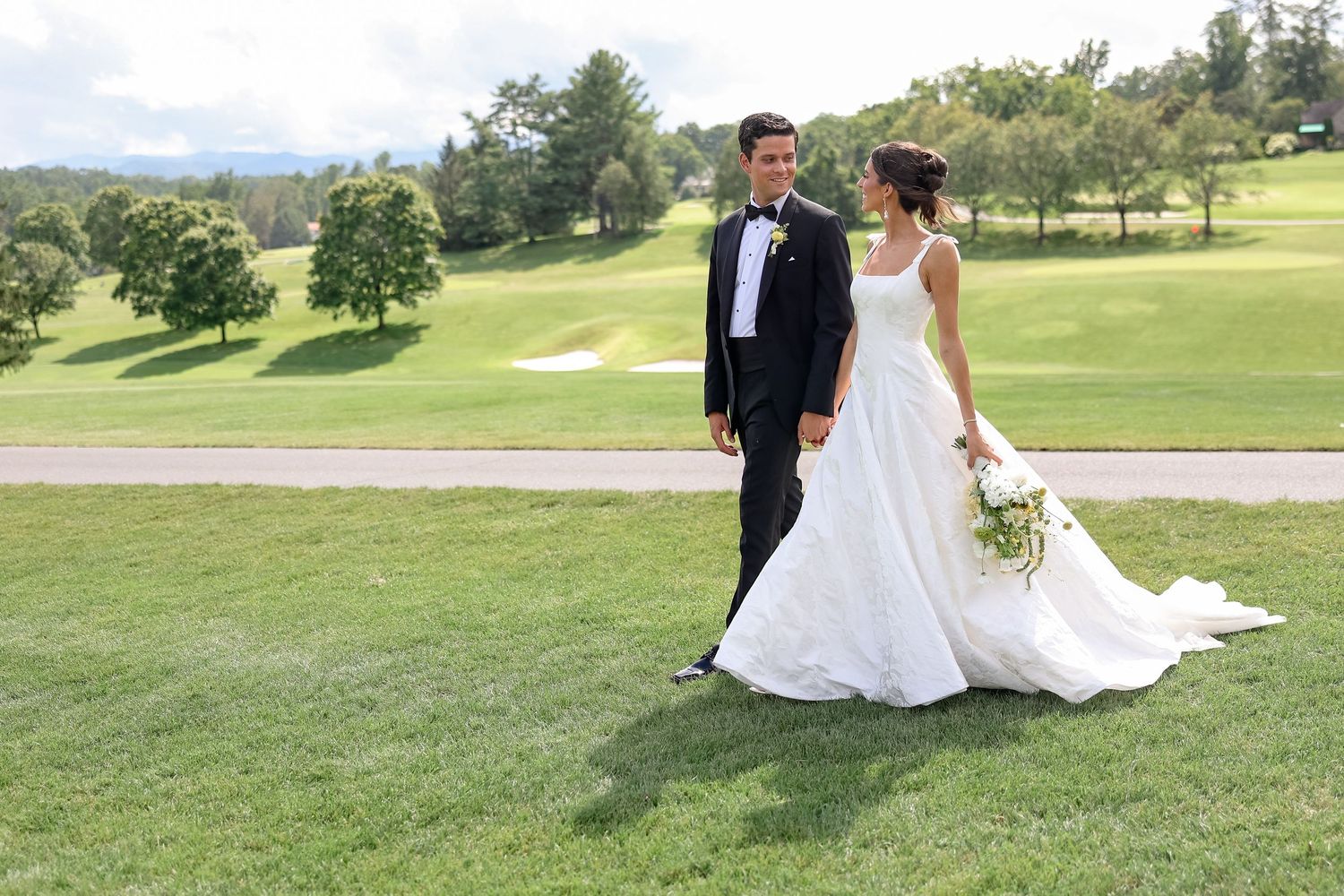 Bride and groom walking on lush green golf course, bride in elegant white gown holding bouquet, groom in classic black tuxedo.
