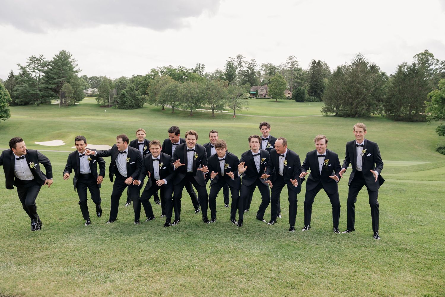 Groomsmen in black tuxedos and bow ties strike fun poses on a green golf course during a wedding celebration.