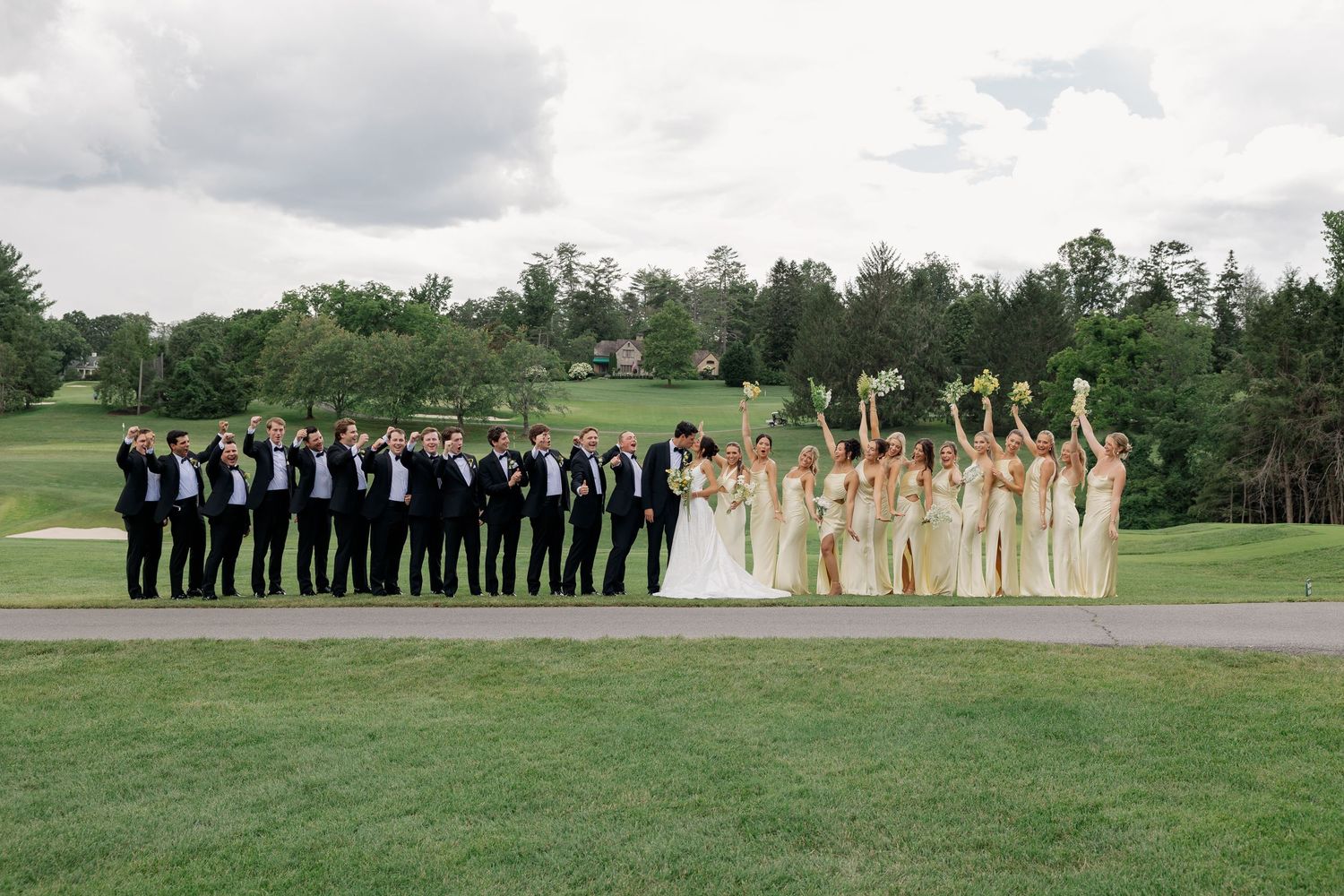 Wedding party poses outdoors, groomsmen in black tuxedos left, bridesmaids in gold dresses right, with bride and groom centered.