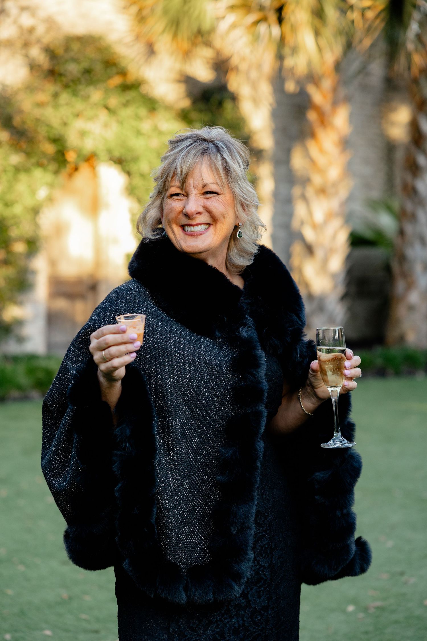 A wedding guest in a black fur-trimmed coat holds champagne glasses while smiling outdoors.