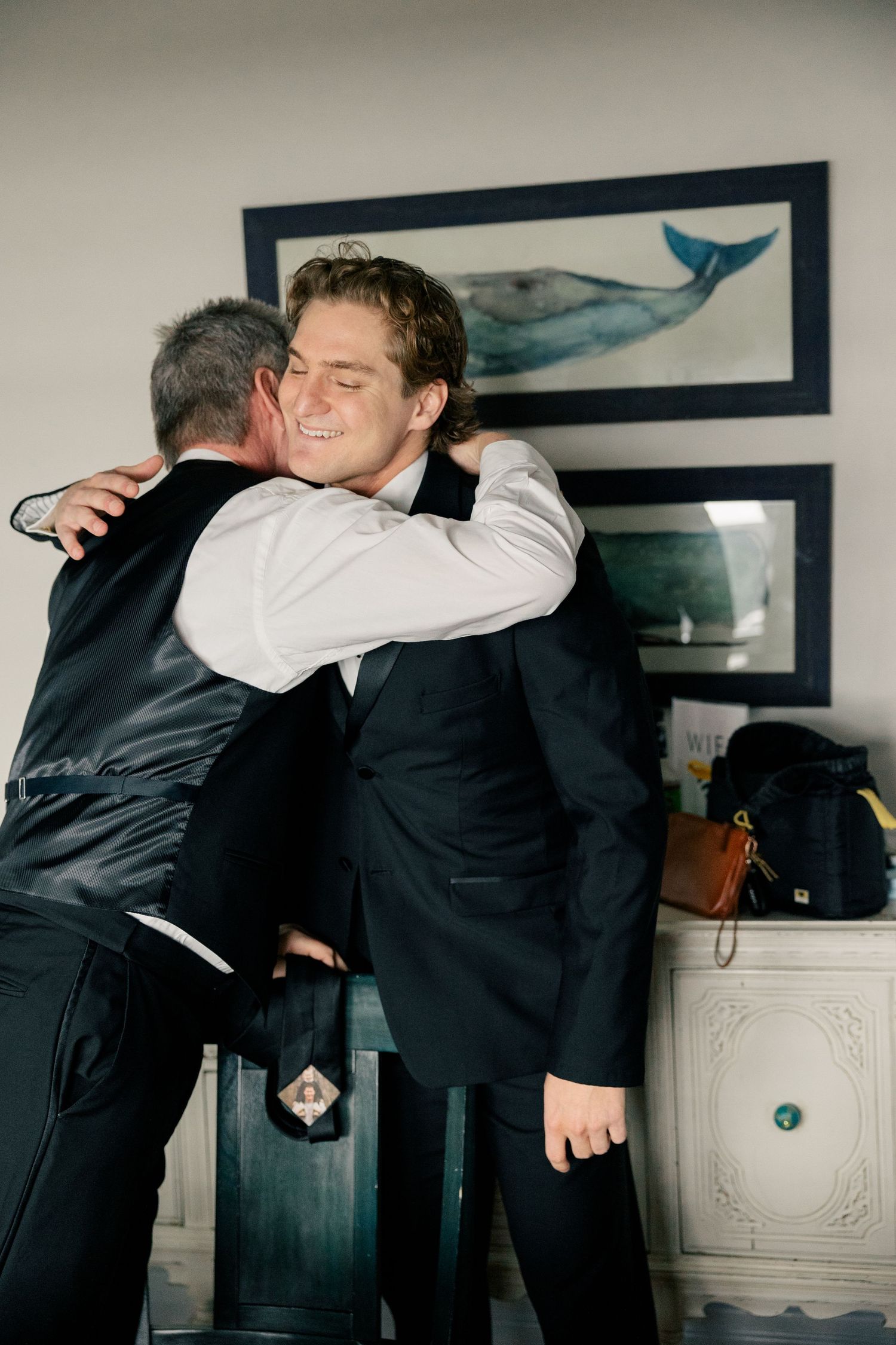 Two people in dark suits share a warm embrace while standing in a room with framed whale artwork on the wall.