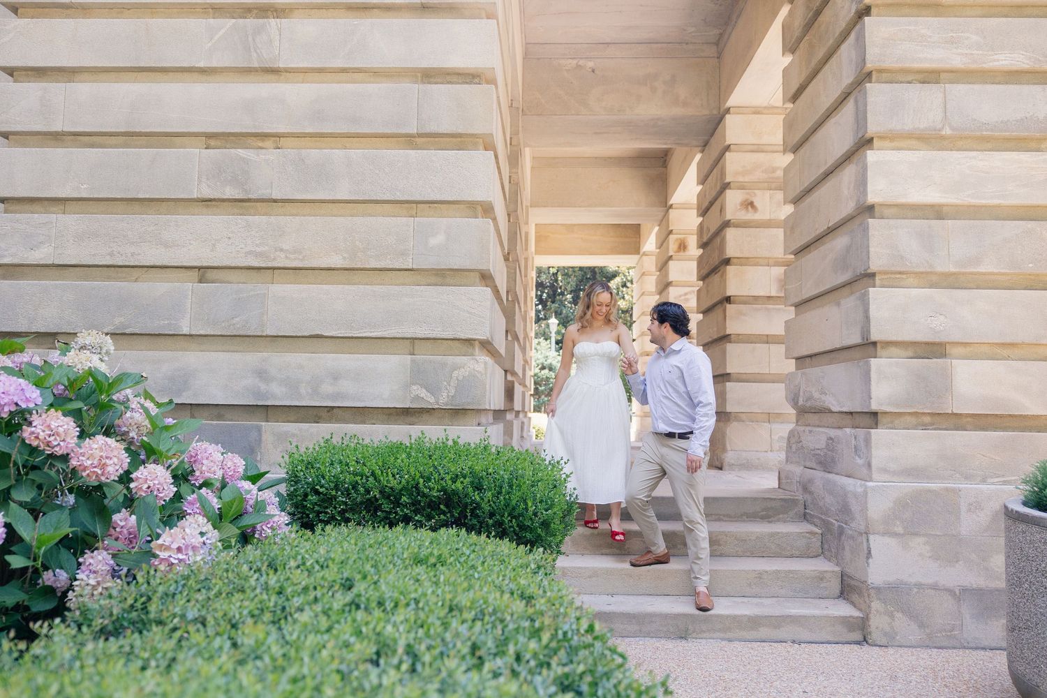 Couple walking hand-in-hand near elegant stone columns surrounded by lush green hedges and pink hydrangeas.