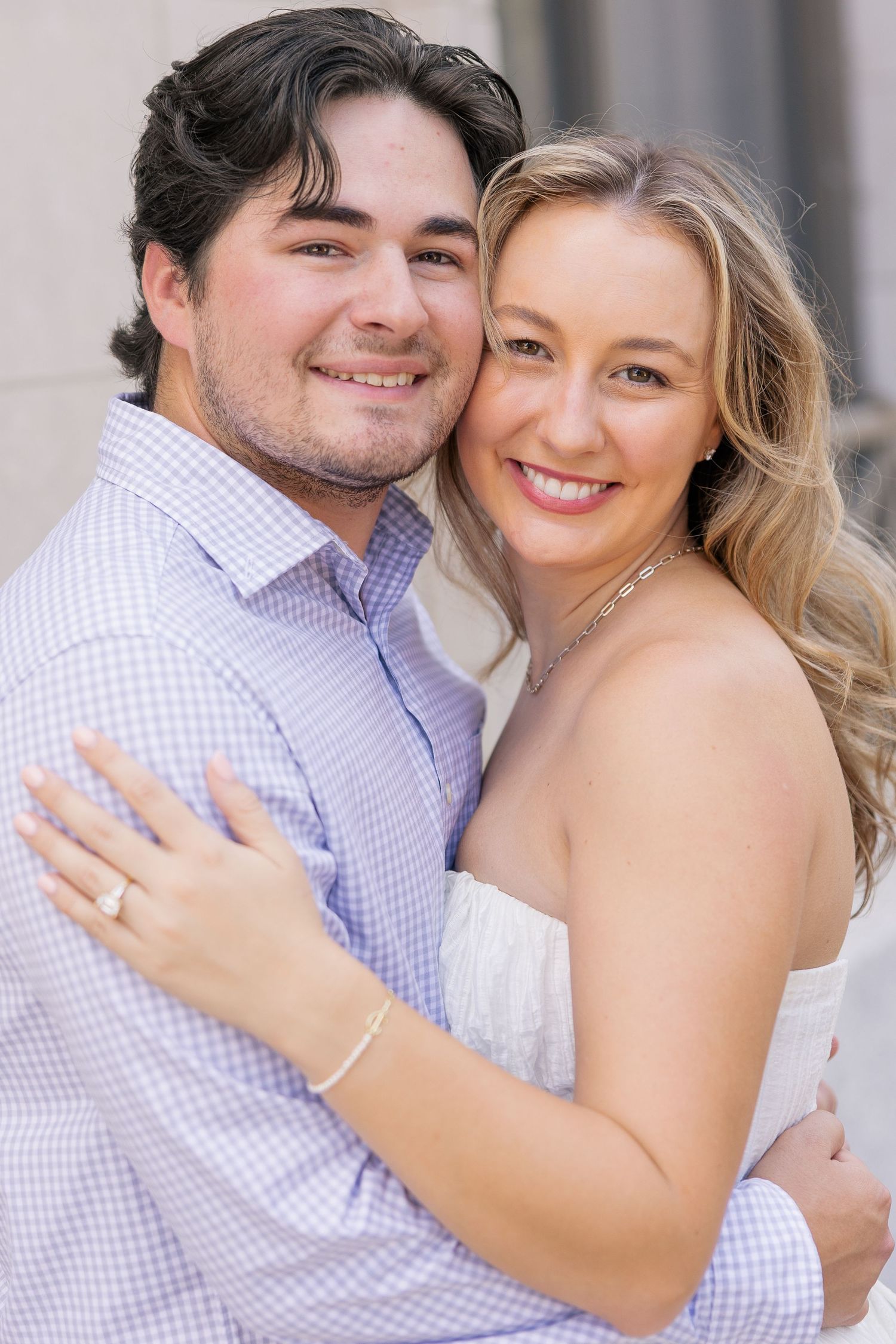 A smiling couple embraces closely, the woman wearing an engagement ring and white dress, the man in a light blue checkered shirt.