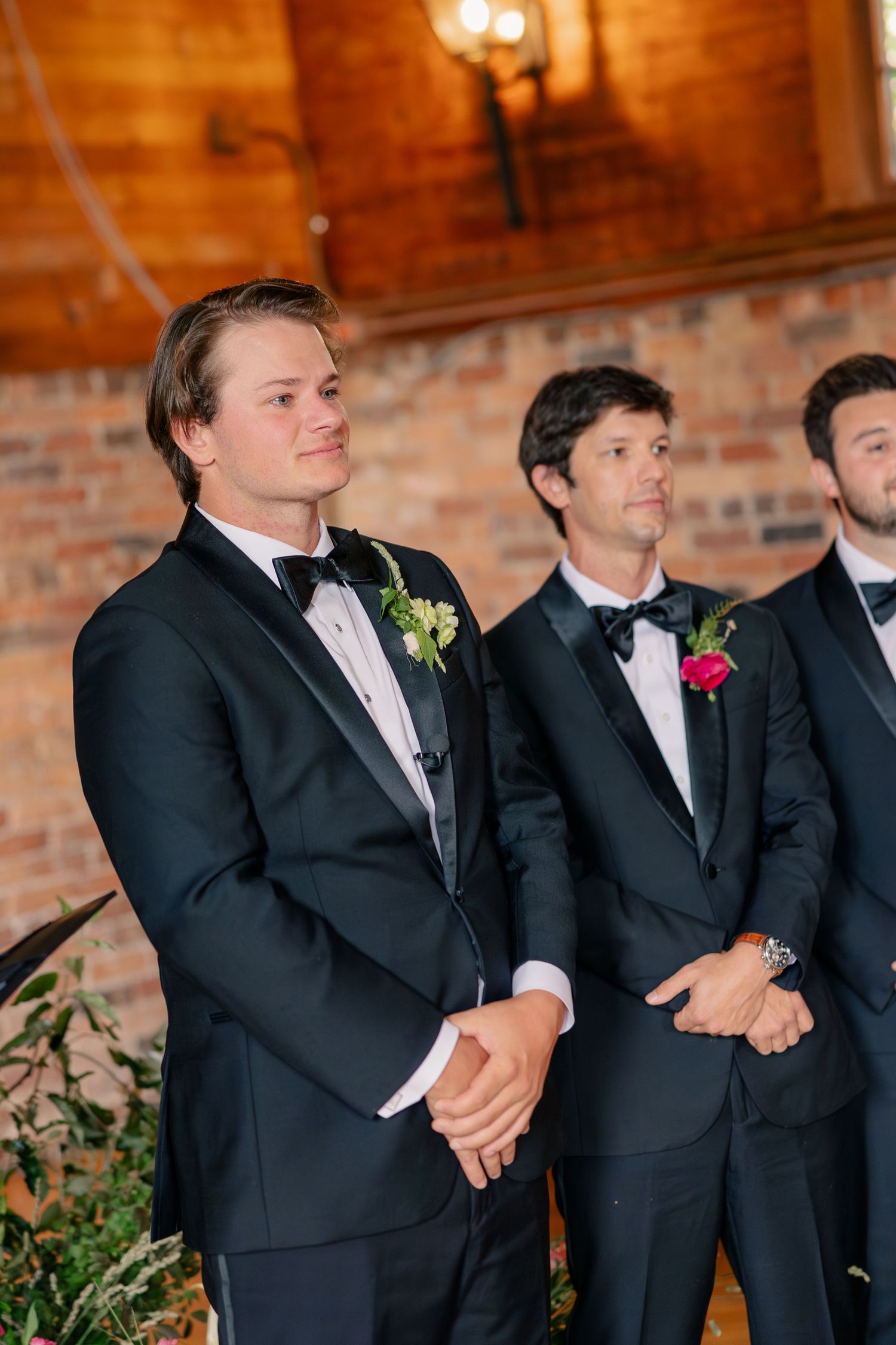 Groomsmen in black tuxedos stand at altar in rustic brick-walled wedding ceremony venue.