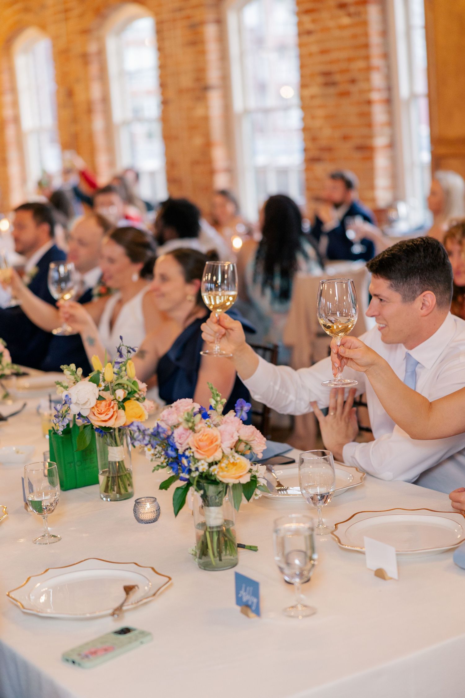 Wedding guests raise glasses of white wine in a toast at a beautifully decorated reception with floral centerpieces.