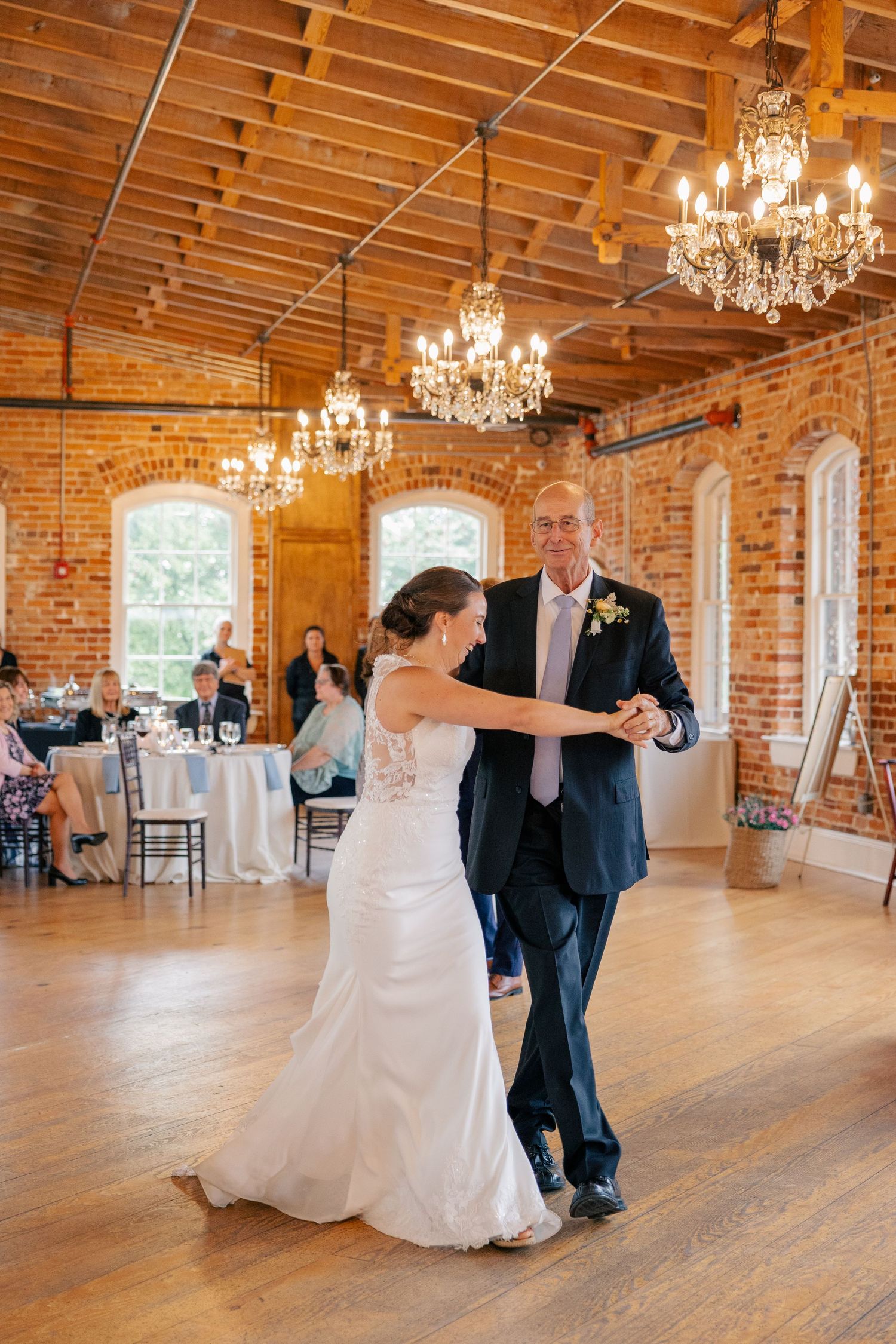 Bride and father share a first dance in an elegant brick-walled reception hall with chandeliers and wooden ceiling.