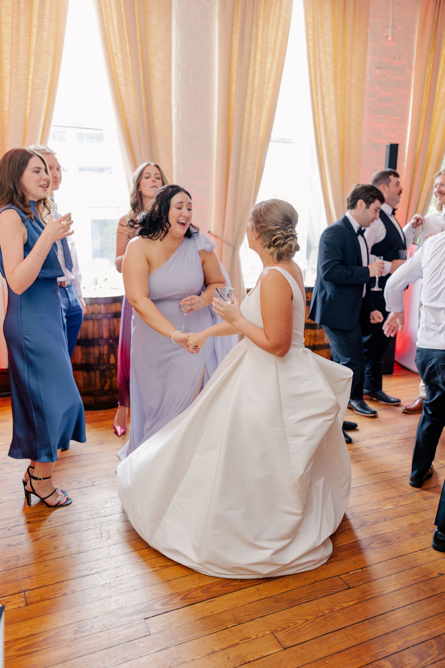 Bride in white gown twirling on dance floor with bridesmaids in blue and lavender dresses at wedding reception.