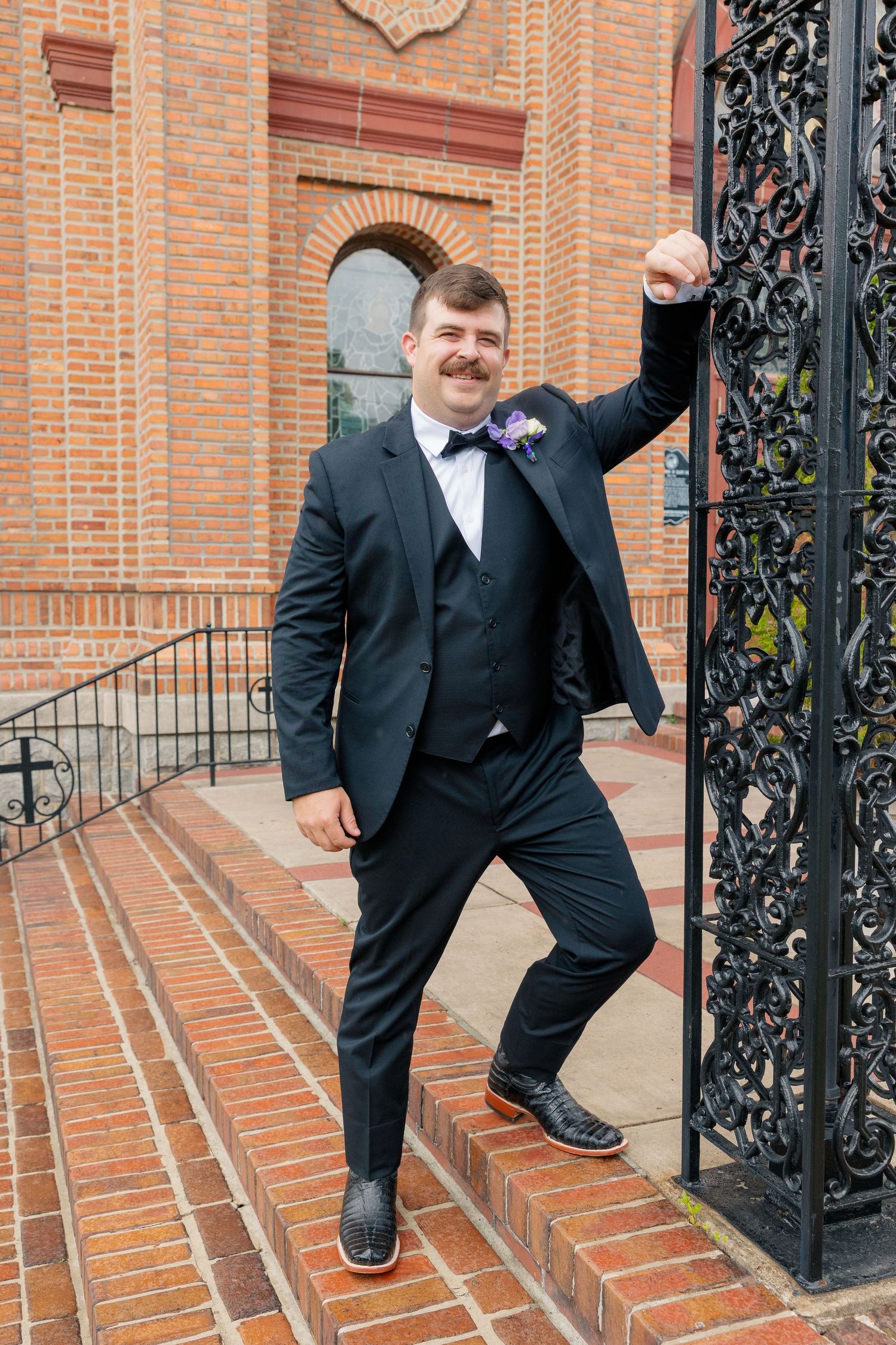 Groom in black tuxedo with purple boutonniere poses at ornate iron gate of brick church.