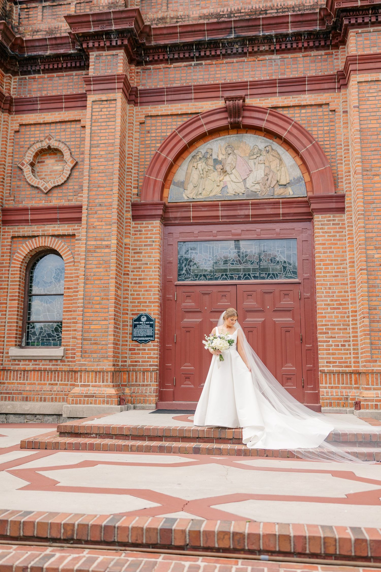 Bride in white gown with cathedral veil stands before grand red brick church entrance.