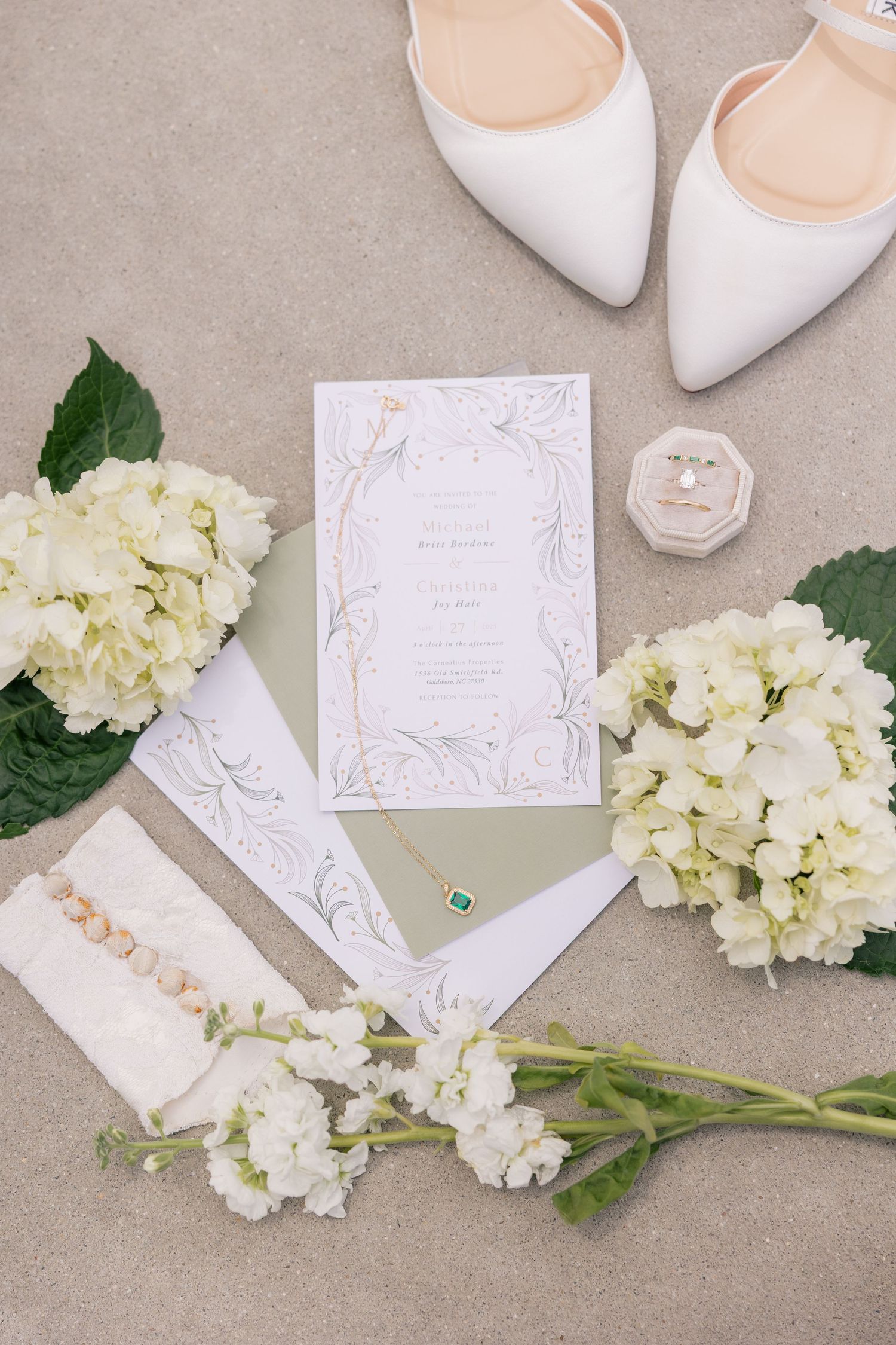 Elegant white bridal accessories including pointed heels, wedding invitations and fresh white hydrangea flowers on beige surface.