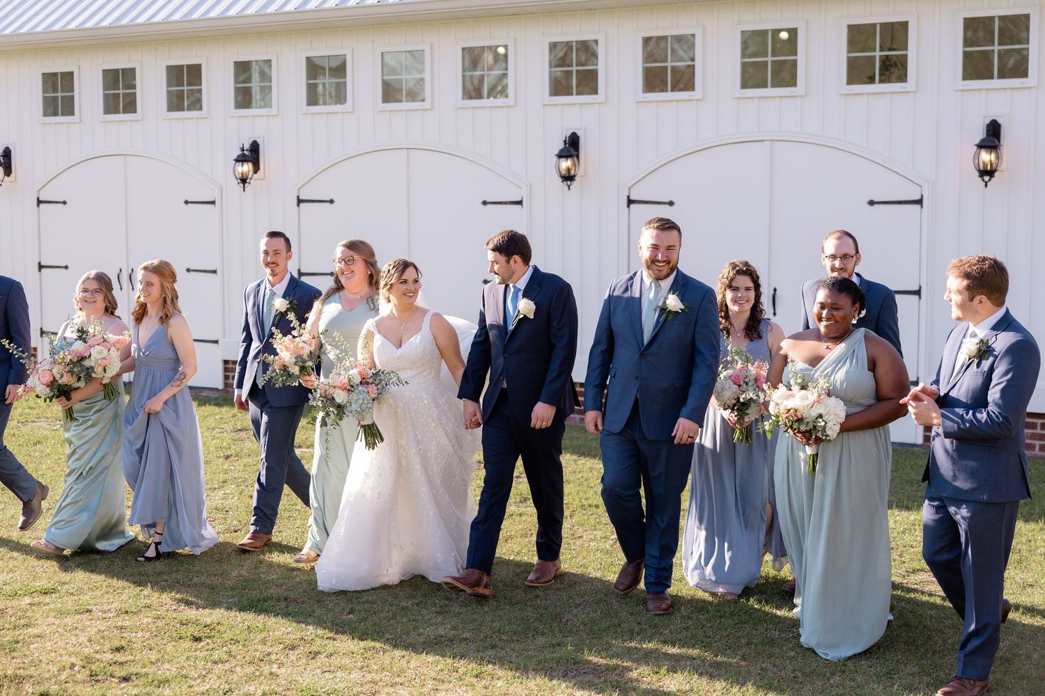 Wedding party poses for photos outside white barn-style venue.