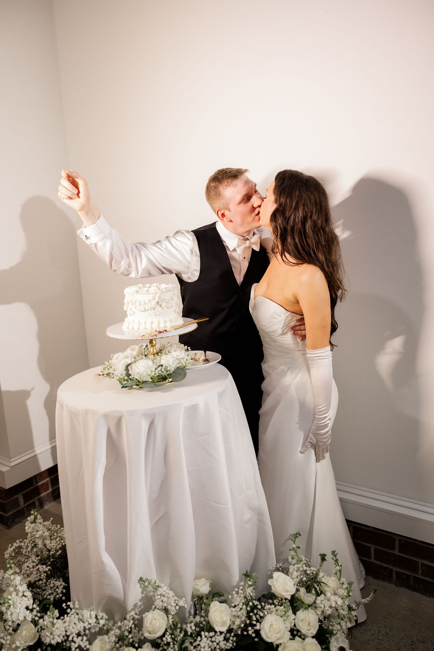 Newlyweds playfully smash wedding cake together at reception against white wall.