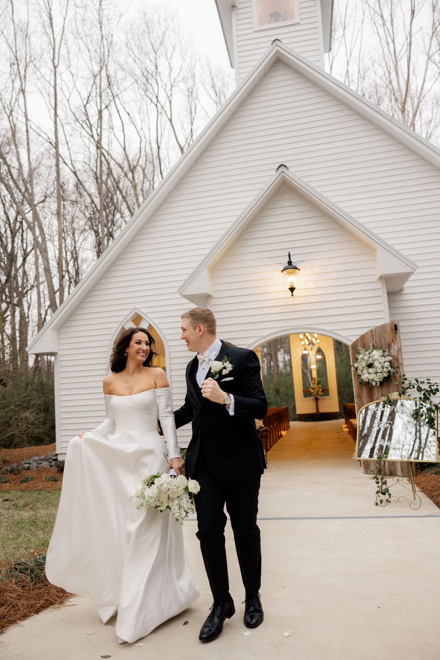 A couple celebrates outside a white chapel wedding venue surrounded by bare winter trees.