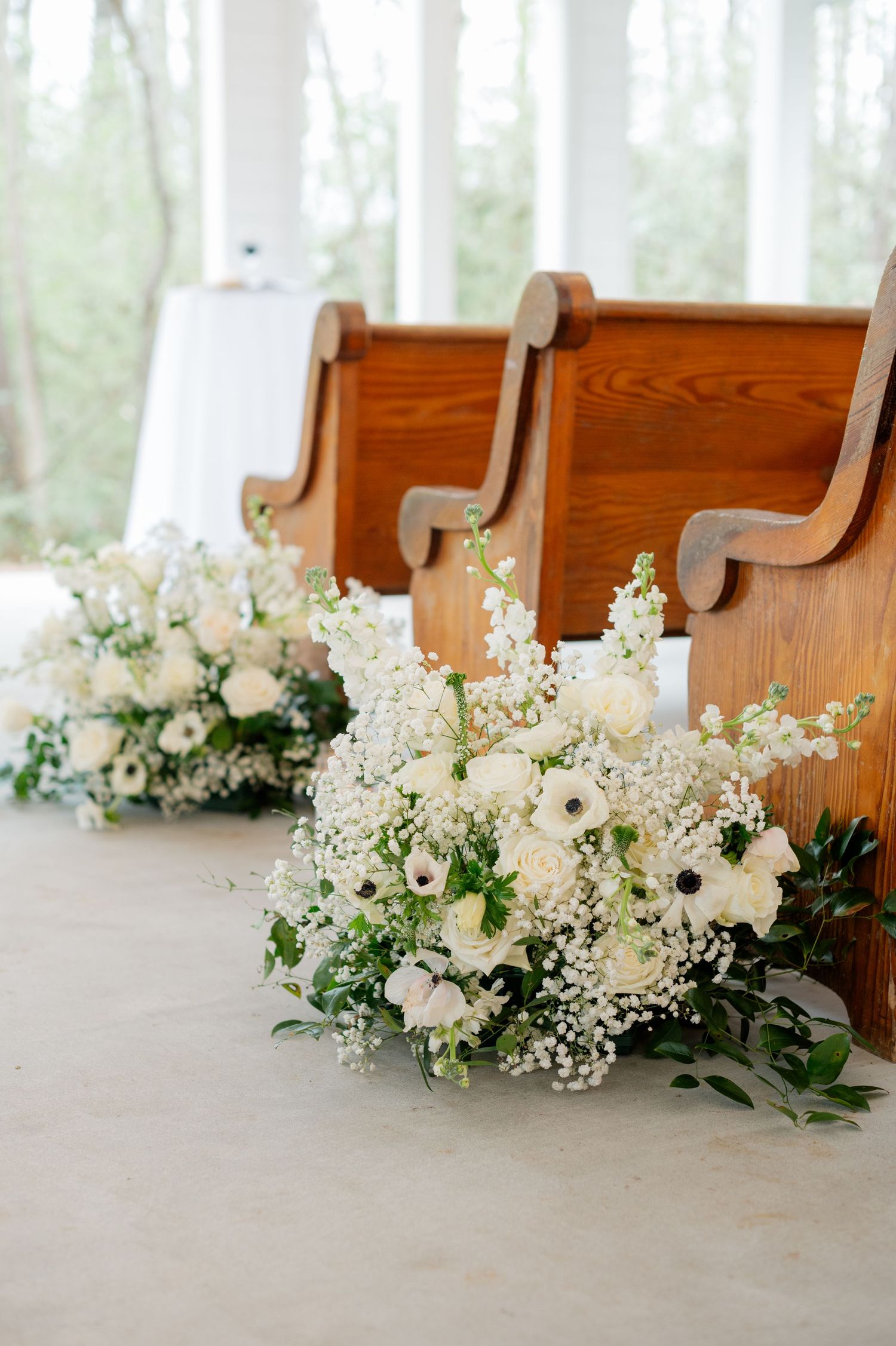 White floral arrangements with delicate baby's breath decorate wooden church pews in a sunlit setting.