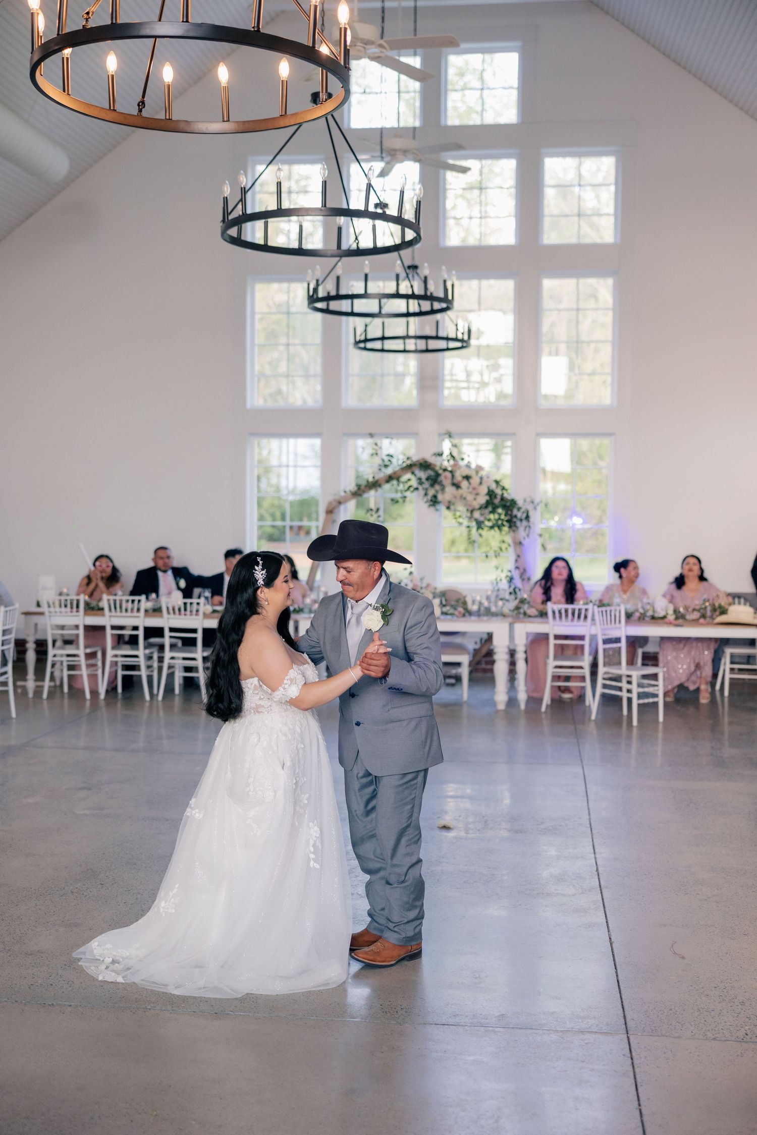 A couple shares a romantic dance moment in an elegant white reception venue with hanging circular chandeliers.