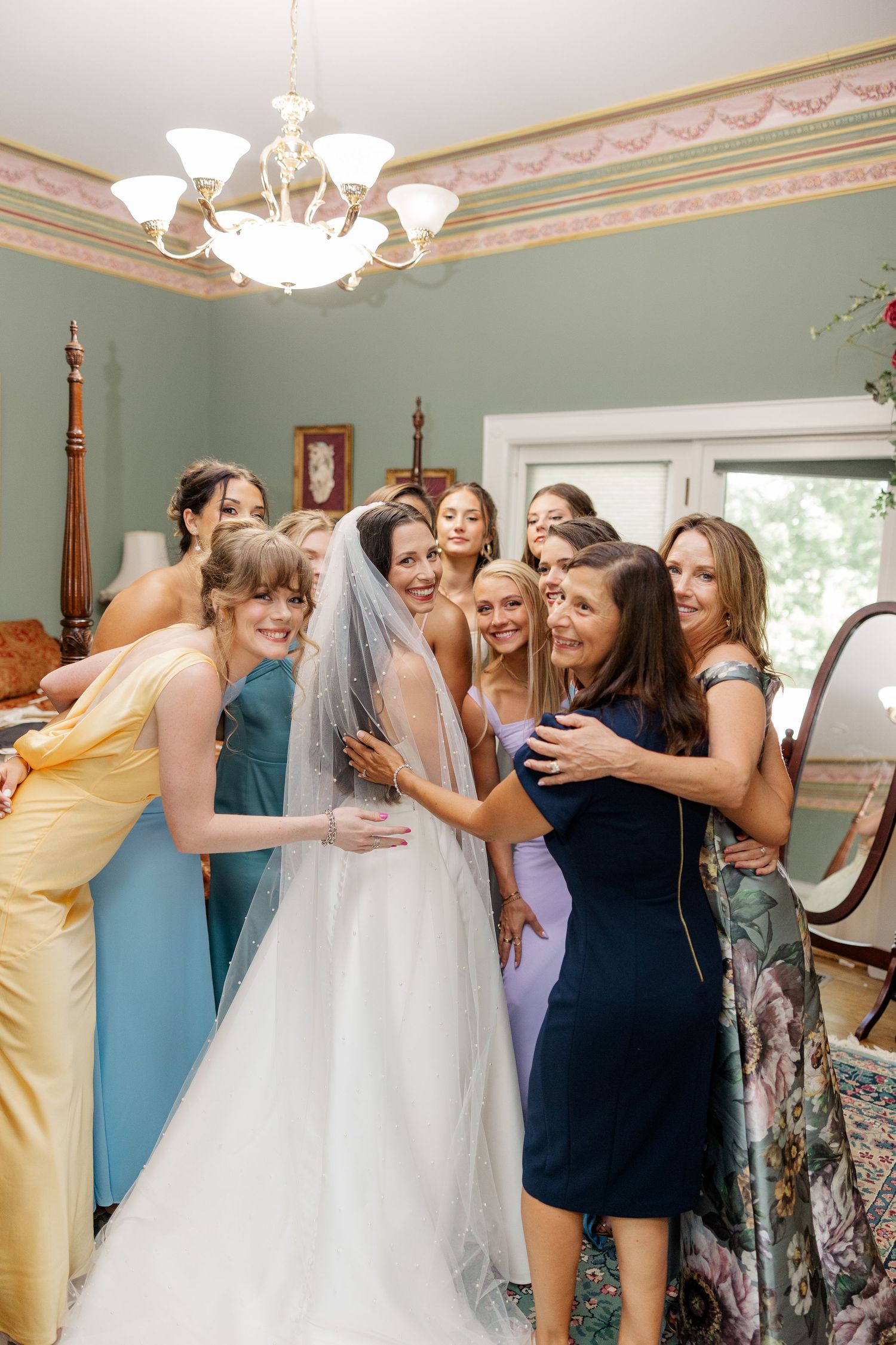 A joyful group celebration scene with bridesmaids in various colored dresses embracing in an elegant room.
