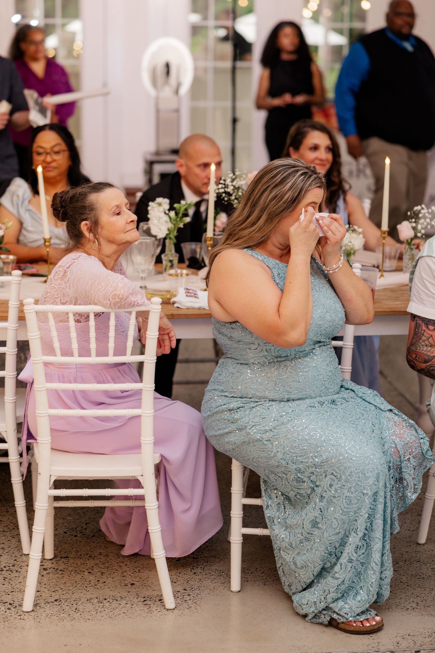 Wedding reception guests in blue dresses seated at tables with white chairs in an elegant indoor venue.