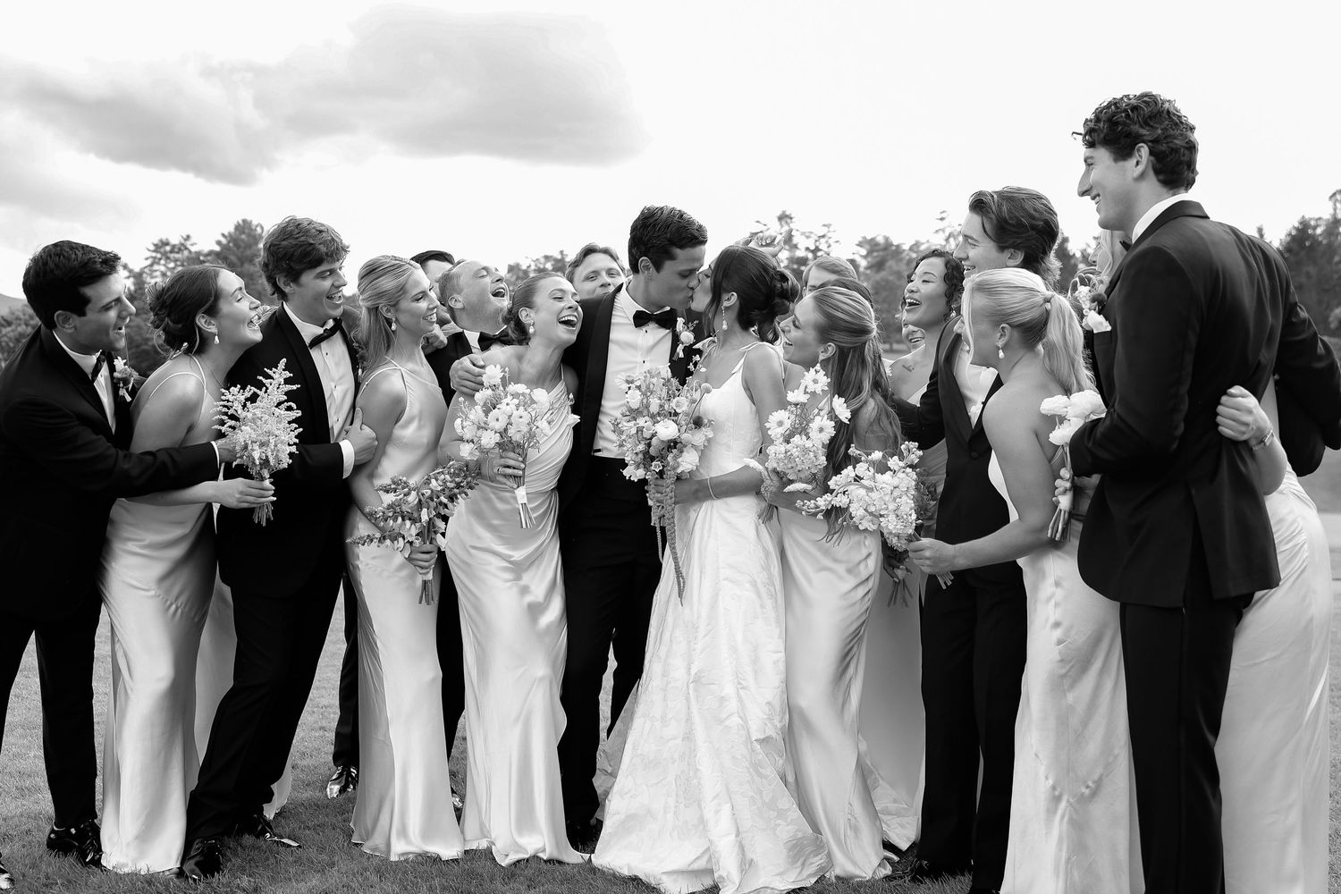Black and white wedding party photo with bride, groom, bridesmaids in gowns and groomsmen in tuxedos celebrating outdoors.