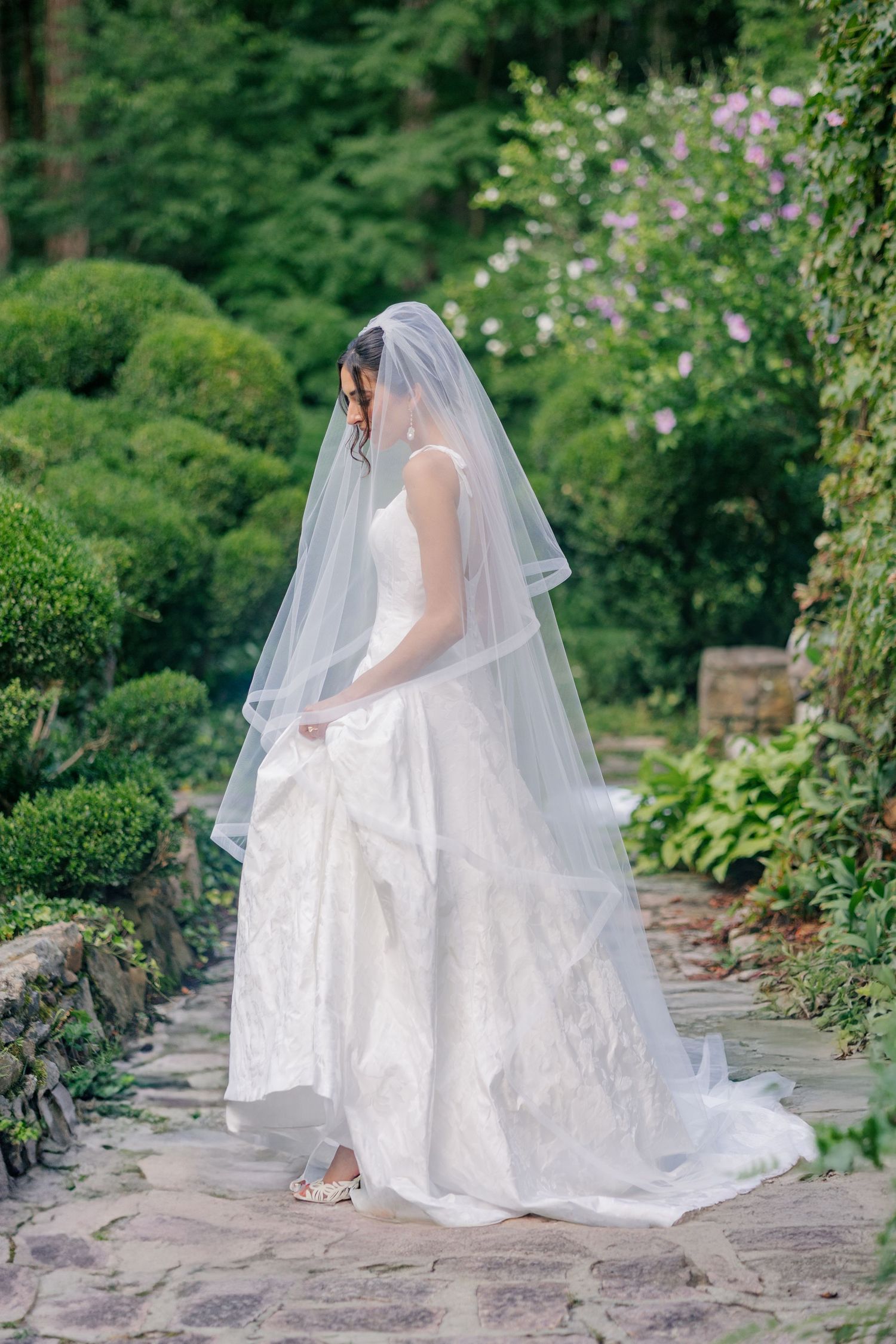 Bride in white lace gown and veil stands on stone path in lush garden with blooming flowers.