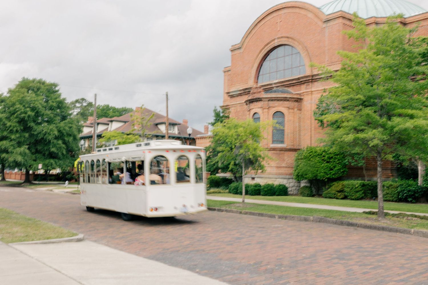 White vintage trolley passing a red brick church on a cobblestone street lined with green trees.