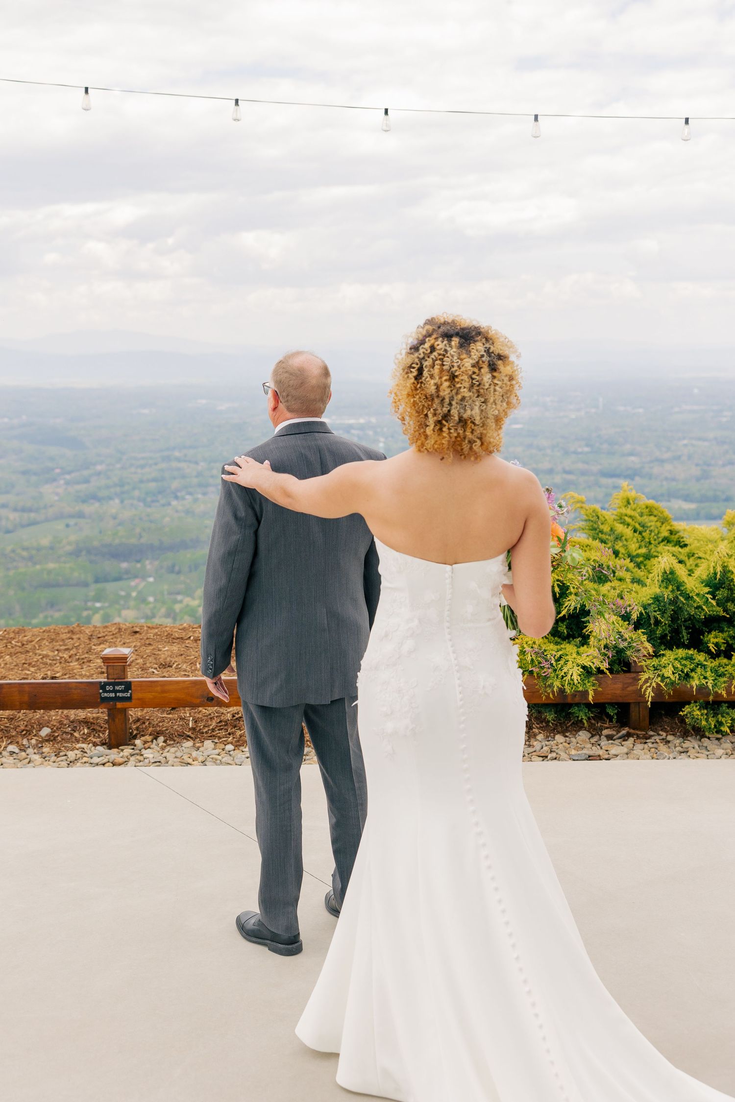 Bride in white strapless gown places hand on groom's shoulder as couple admires panoramic landscape view.