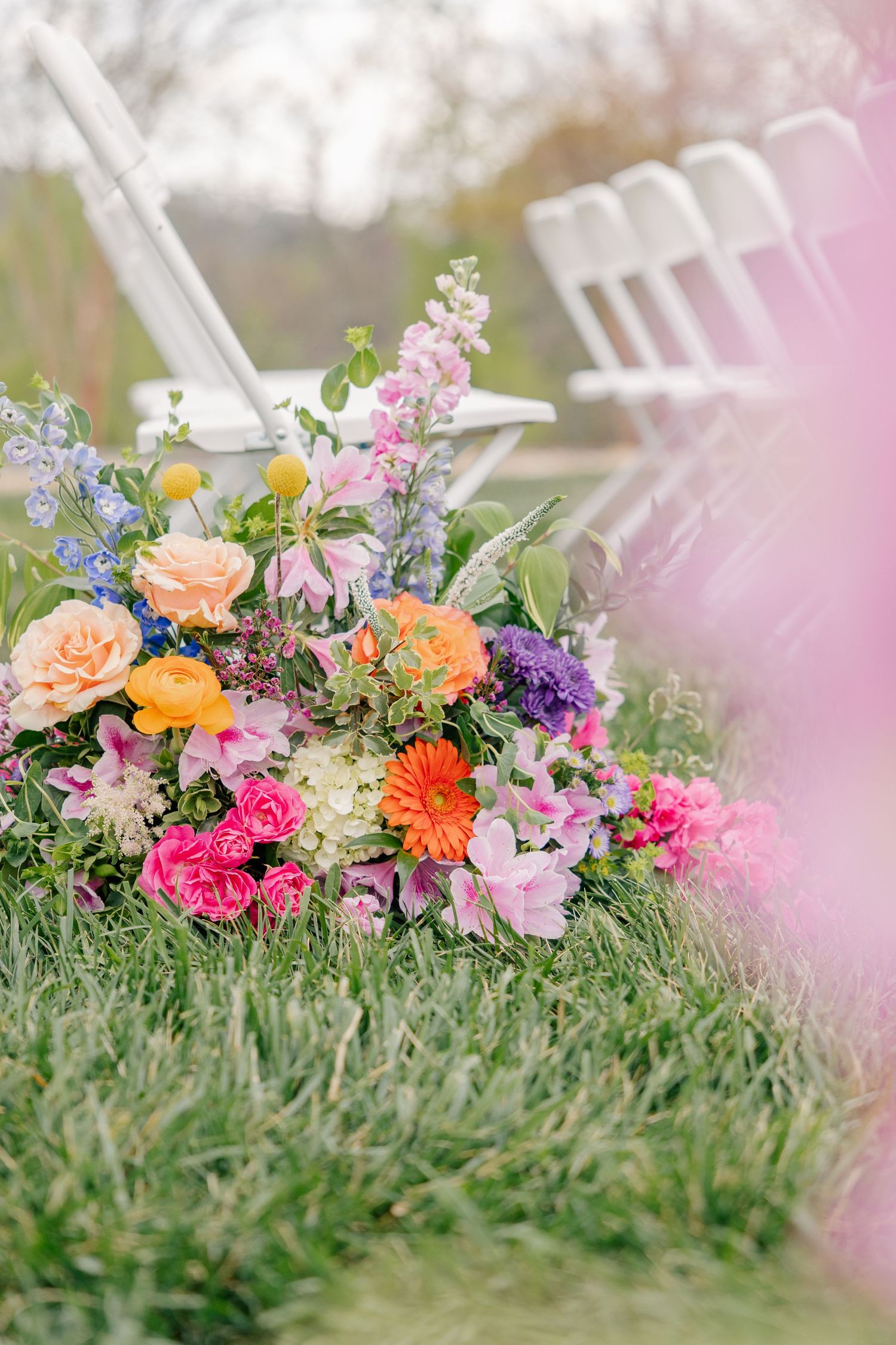Colorful wedding ceremony floral arrangement on grass beside white chairs with pink, orange, and purple flowers.