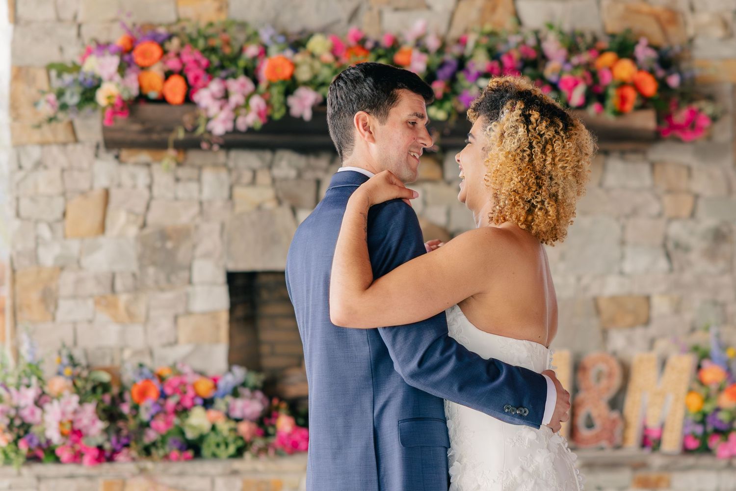 Bride and groom share romantic moment in front of stone fireplace adorned with colorful floral arrangements.