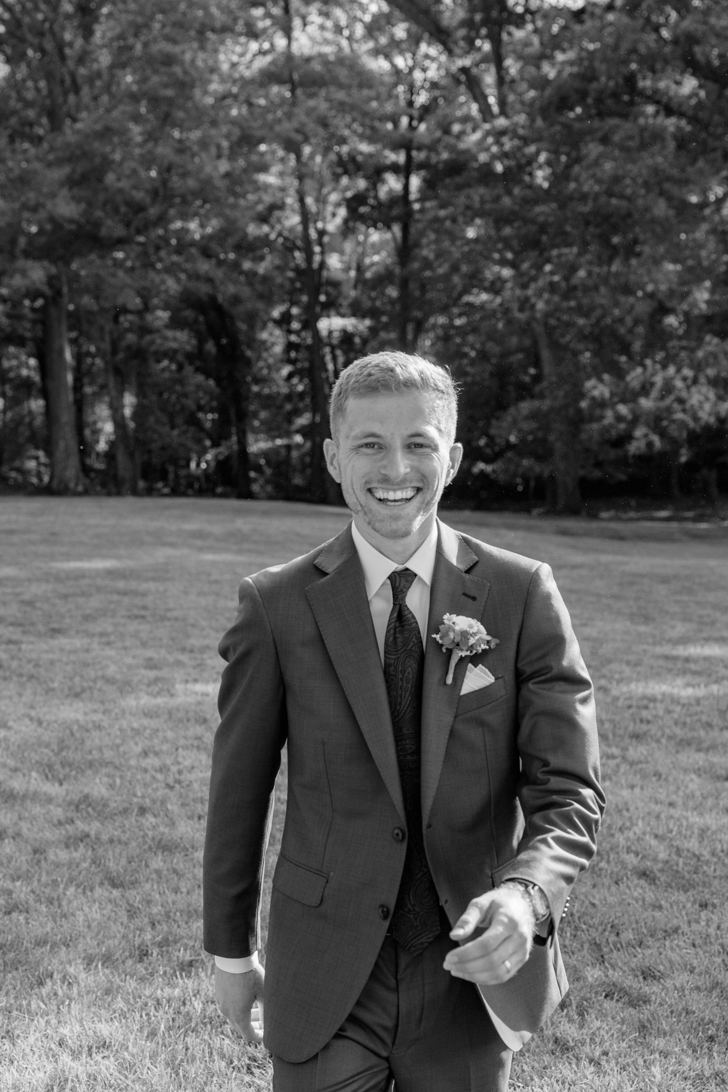 Smiling groom in dark suit with boutonniere poses outdoors on green lawn with trees in black and white photo.