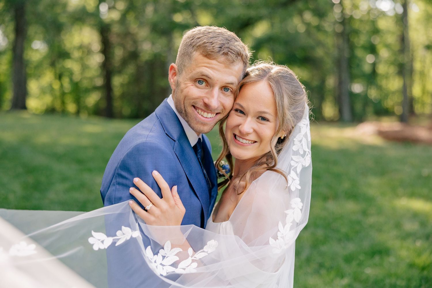 Smiling bride and groom embrace outdoors with flowing lace veil in sunlit green setting.